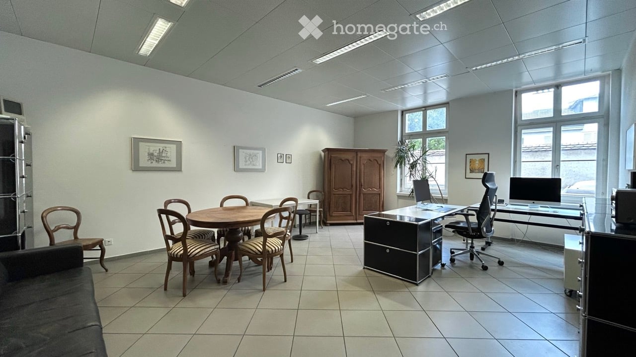Bright, modern office space with a mix of furniture including a sofa, round wooden table with chairs, office desk with computer, and a large cabinet. The room features large windows, tiled flooring, and several framed pictures on the walls.