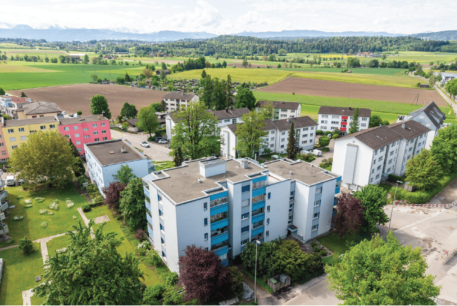 Aerial view of a residential area with multiple apartment buildings, surrounded by green spaces, trees, and a scenic mountain backdrop.