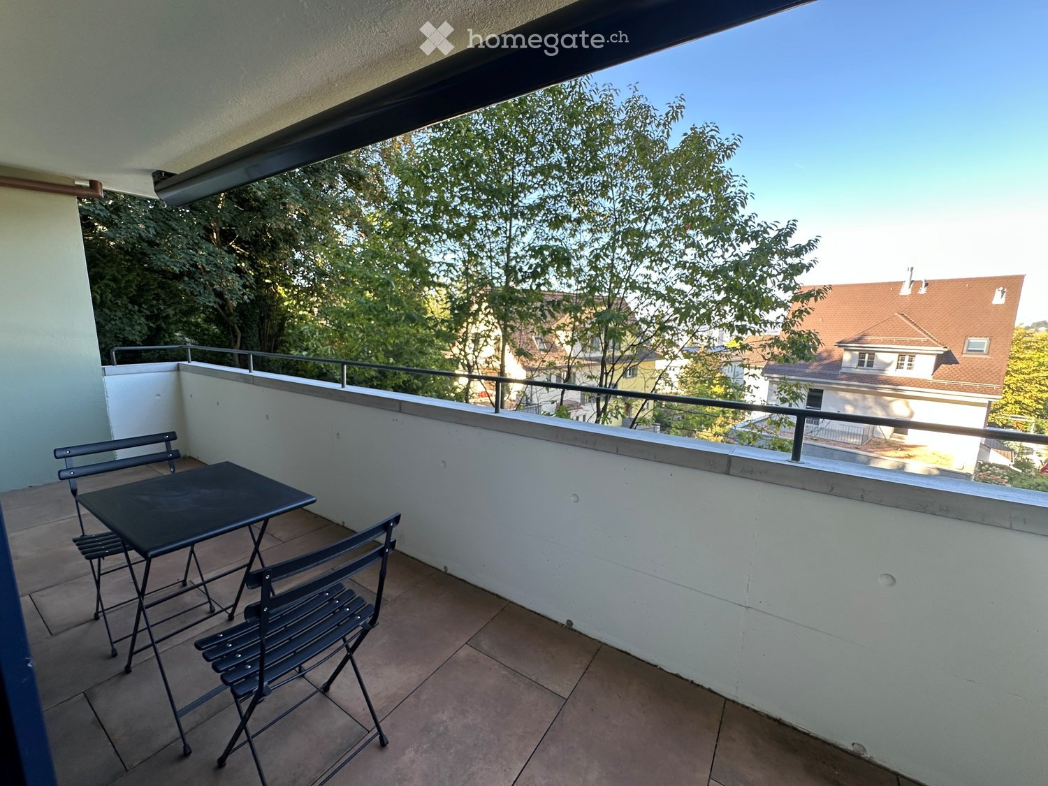 Balcony with tiled floor, white walls, metal table and chairs, metal railing, and view of trees and houses.
