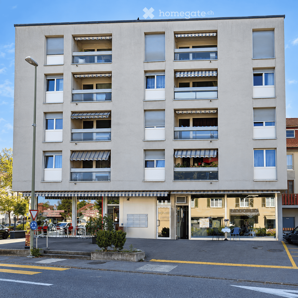 Four-story residential building with multiple balconies, striped awnings, and a storefront at the ground level. Windows have white frames and curtains.