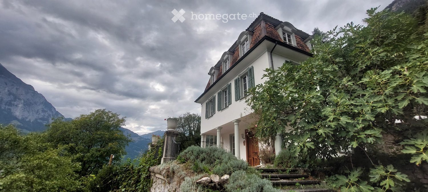 A two-story white house with a red-tiled roof, surrounded by lush greenery and mountains in the background.