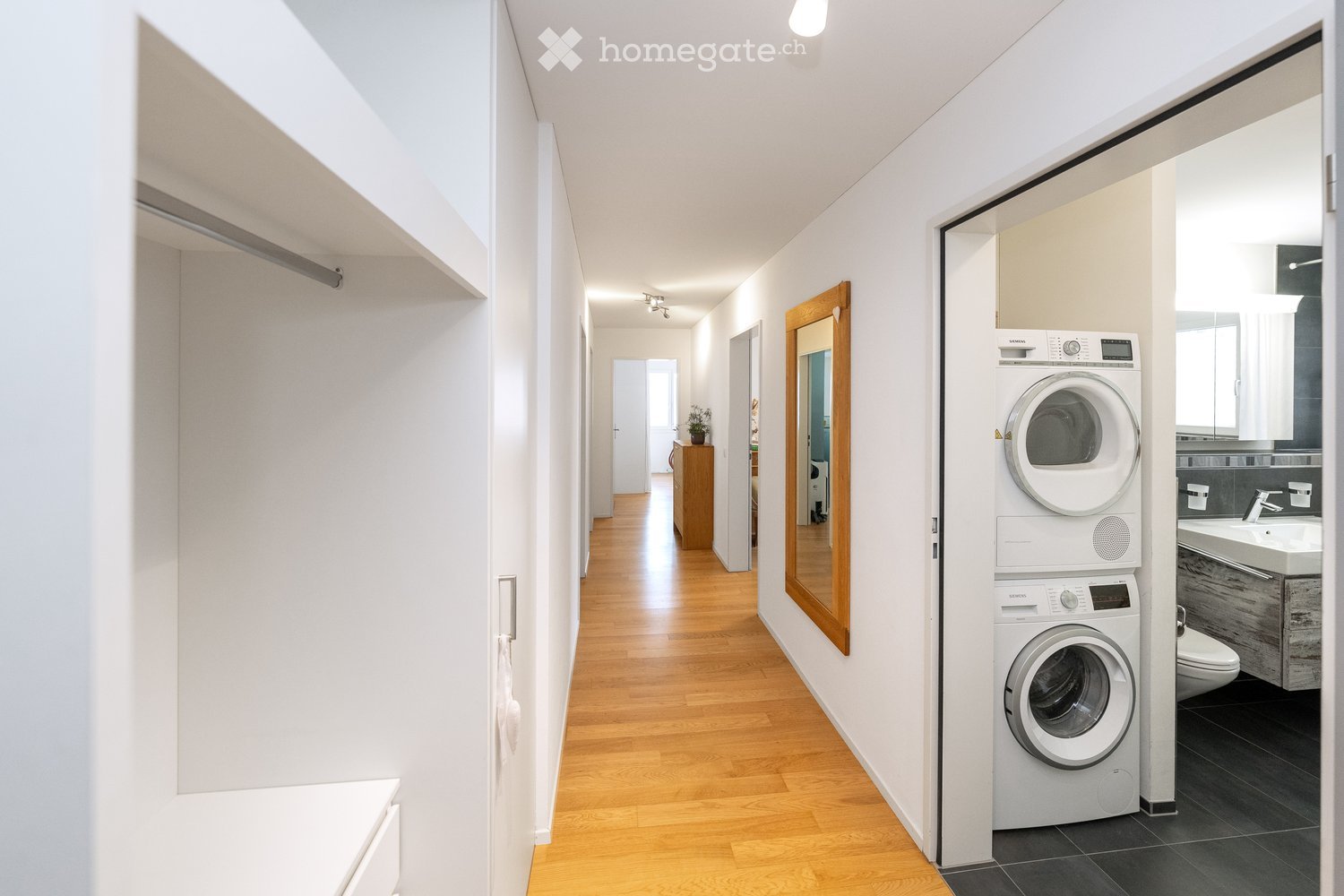 Bright hallway with wooden flooring, white walls, and a private washing machine in the corner. A small cabinet and a mirror are visible further down the hallway.