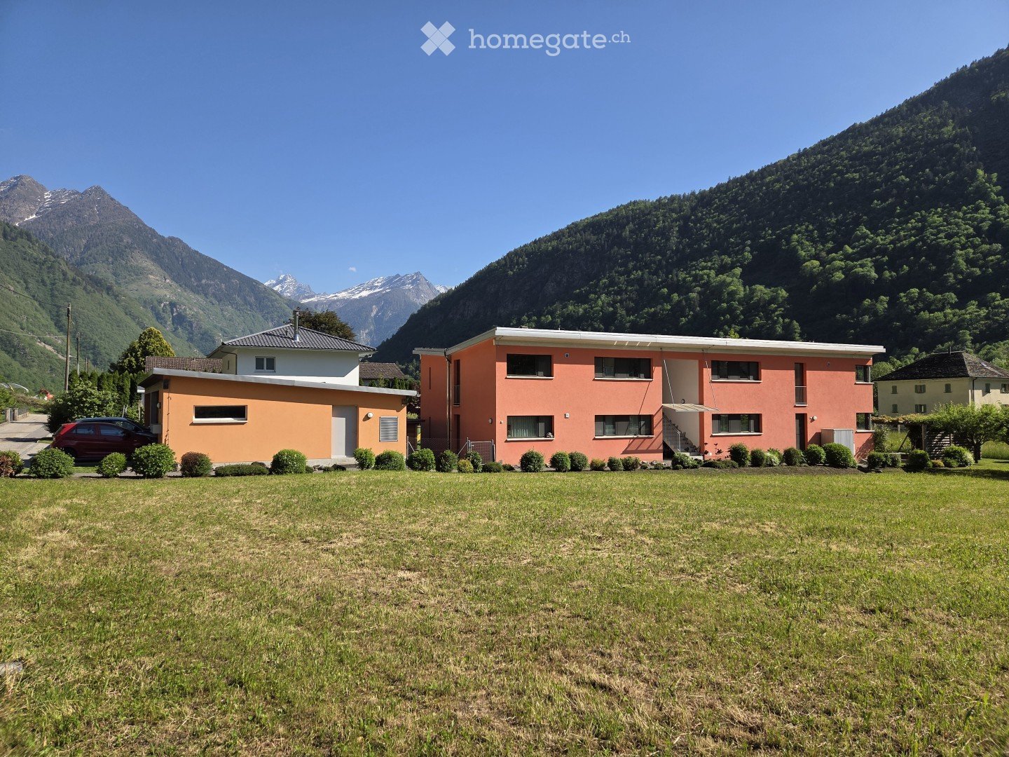 Two-story house with orange and white exterior, surrounded by a grassy lawn and small bushes, with mountains in the background.
