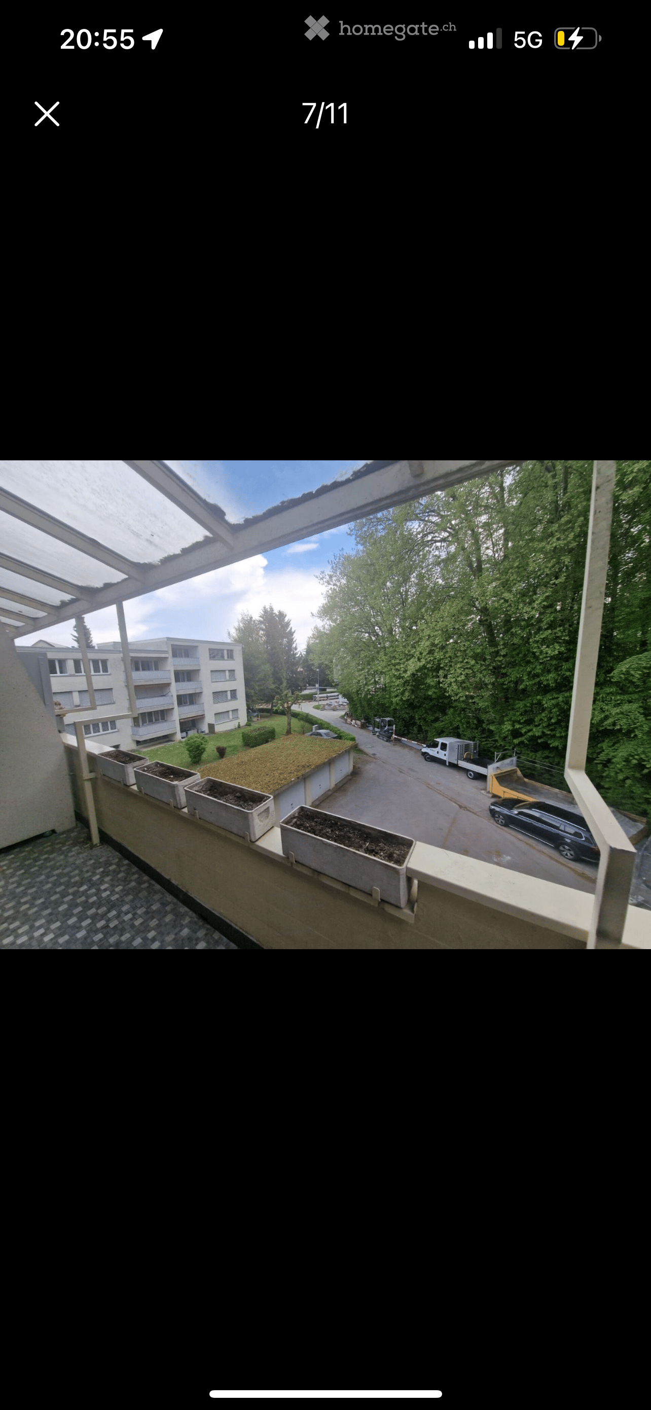 Balcony of an apartment building, with concrete planters, metal railing, asphalt covered ground, green bushes, trees, multi story building, road, parked cars.