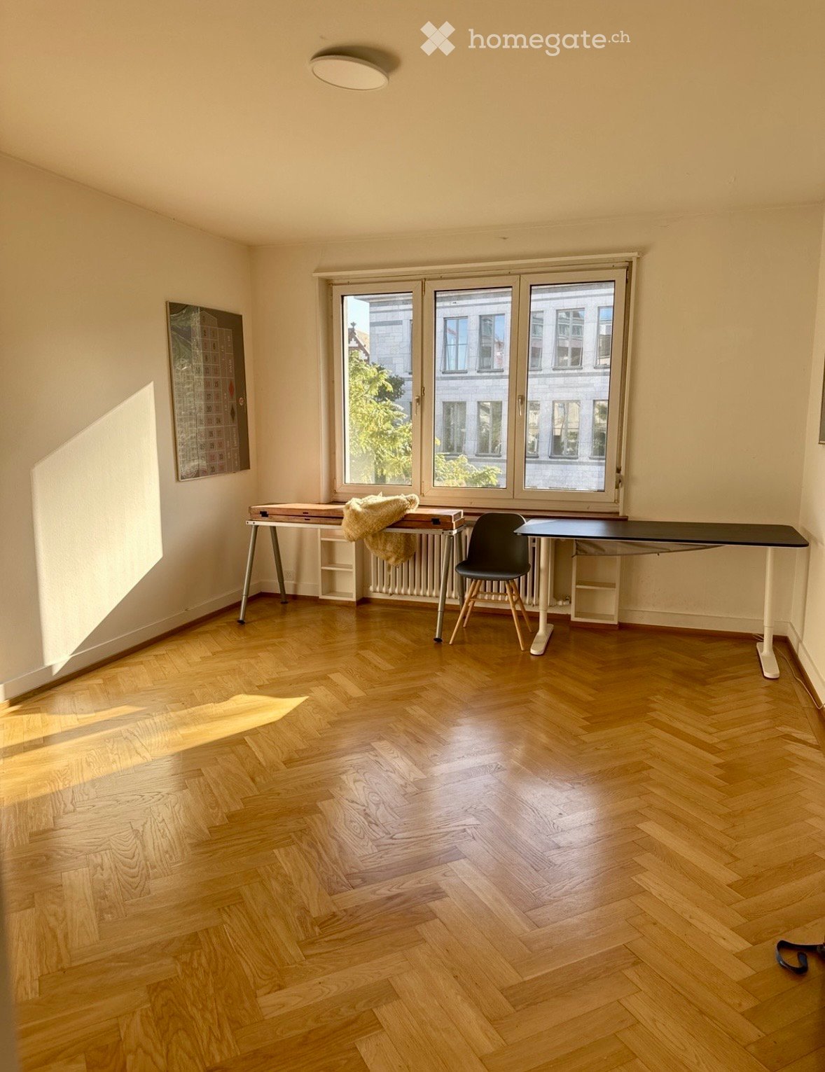 Bright room with wooden herringbone flooring, large windows, a table with a chair, a radiator, and a picture frame on the wall.