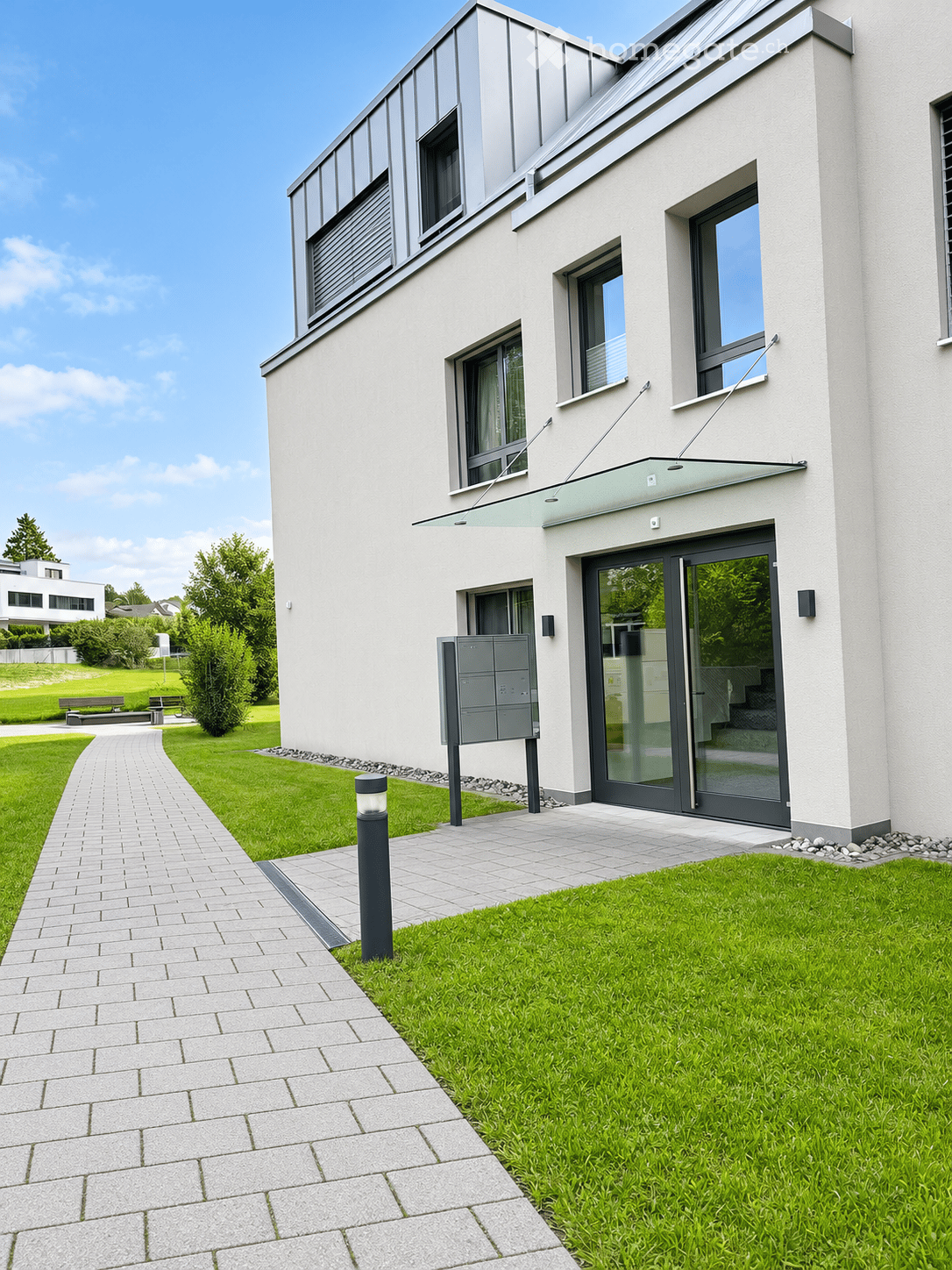 Modern two-story house with white facade, large windows, and a sleek entrance with glass doors. The surrounding area features a well-maintained lawn, a paved walkway, and a mailbox.