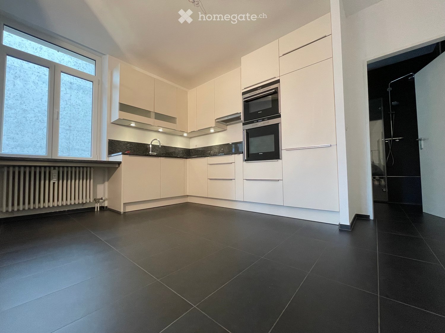 Modern kitchen with white cabinetry, black tiled floor, stainless steel oven, and a sink with a faucet. The space is bright with large windows and a radiator under the window.