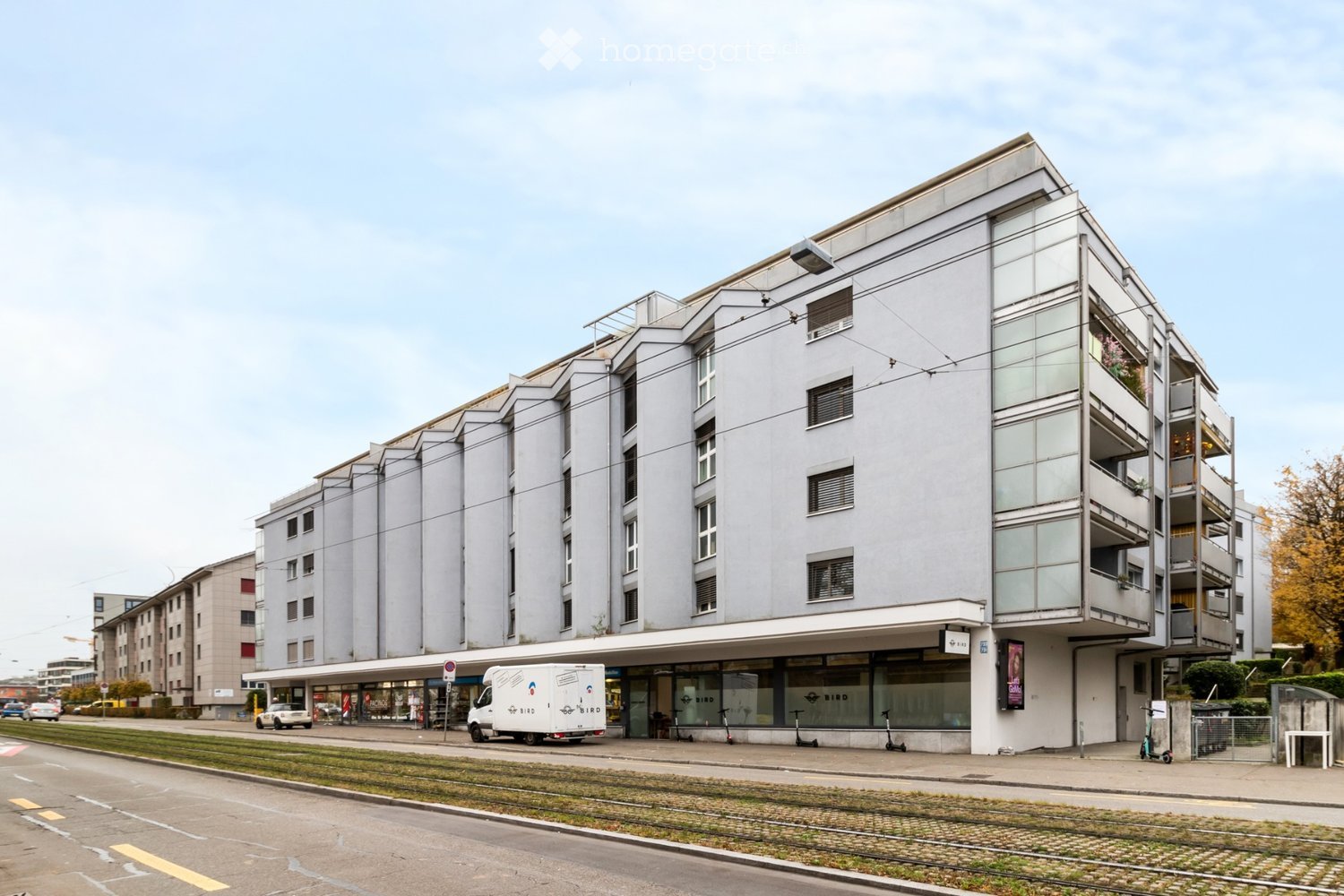 Modern multi-story building with a combination of glass and concrete facade, featuring balconies and large windows. The ground floor has a storefront with glass doors and a sign, and there are scooters parked outside.