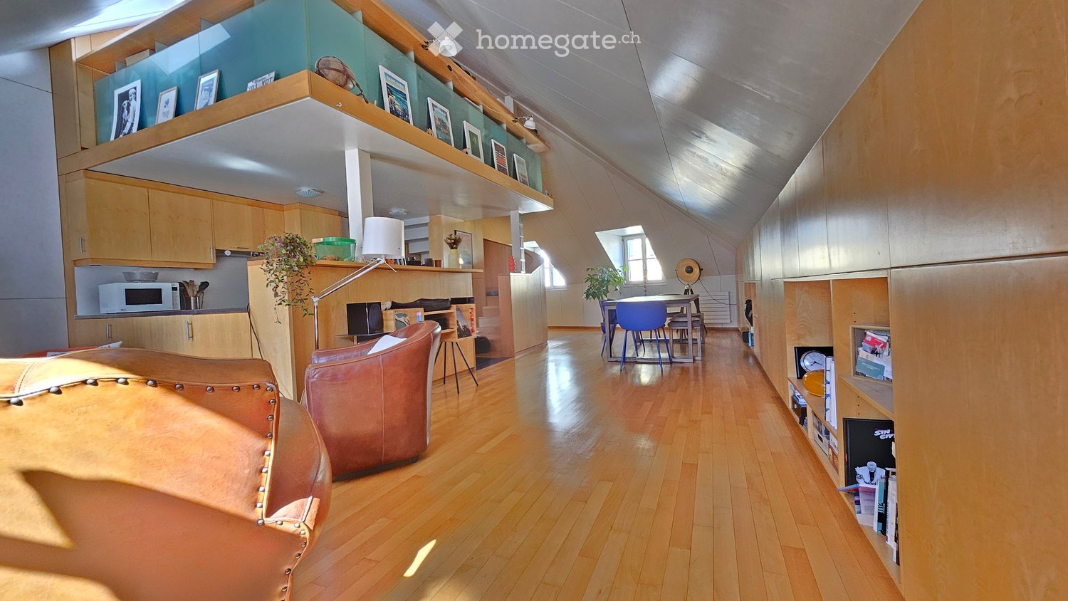 Open-plan living and dining area with wooden flooring, modern furniture, and built-in shelving. Features a leather sofa, dining table with chairs, microwave, and decorative items on shelves.