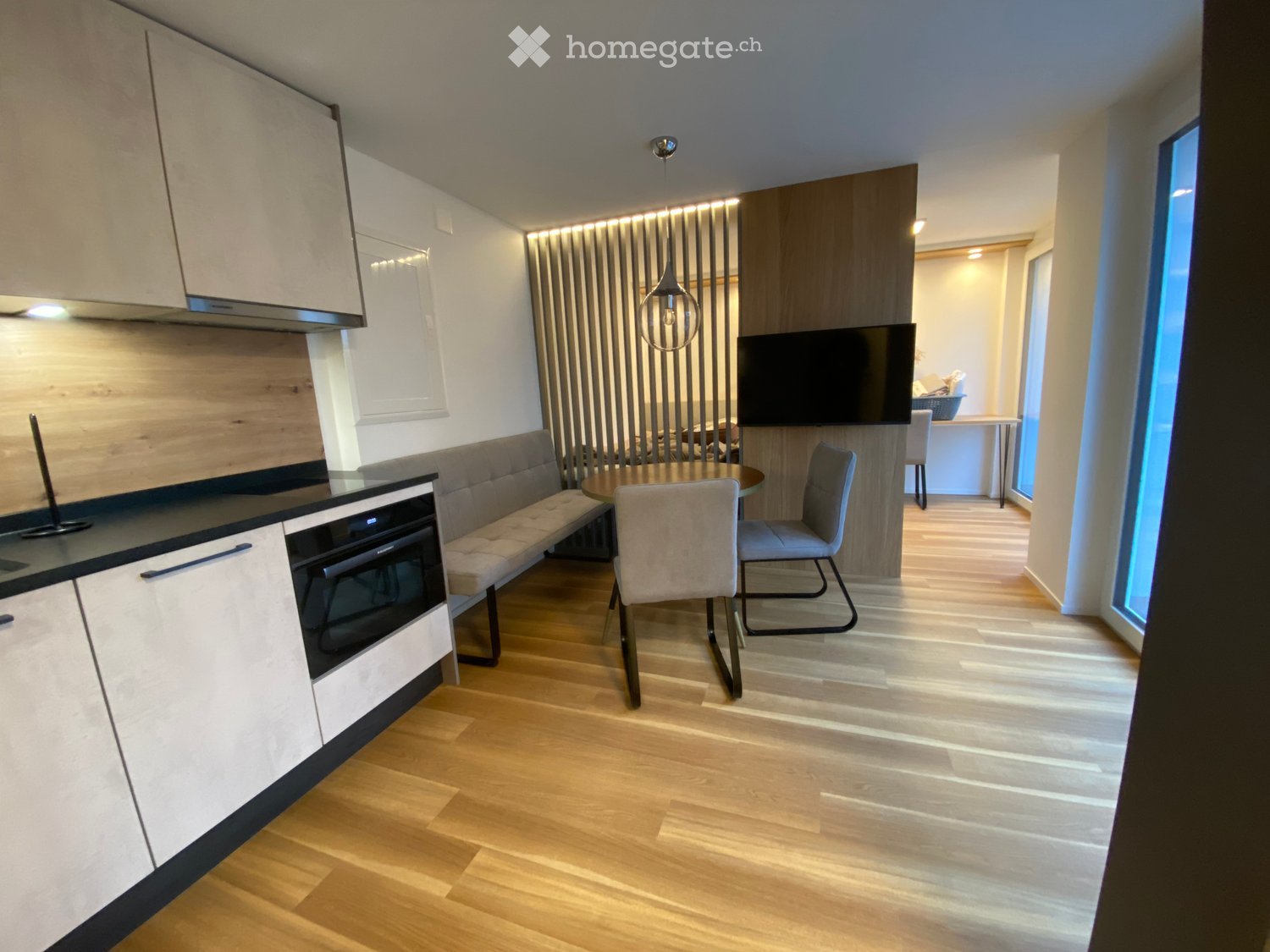 Modern, open-plan living and dining area with light wood flooring, white cabinets, a built-in oven, a bench seating, a dining table with chairs, and a television mounted on the wall.