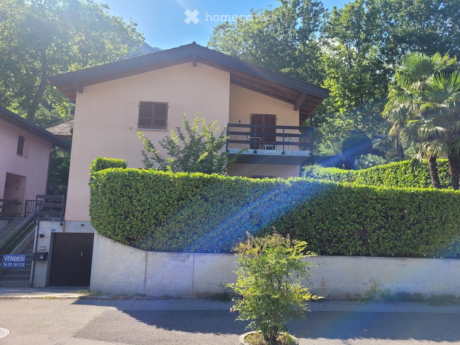 Two-story house with light pink exterior, wooden shutters, and a balcony on the upper level, surrounded by greenery and a hedge.