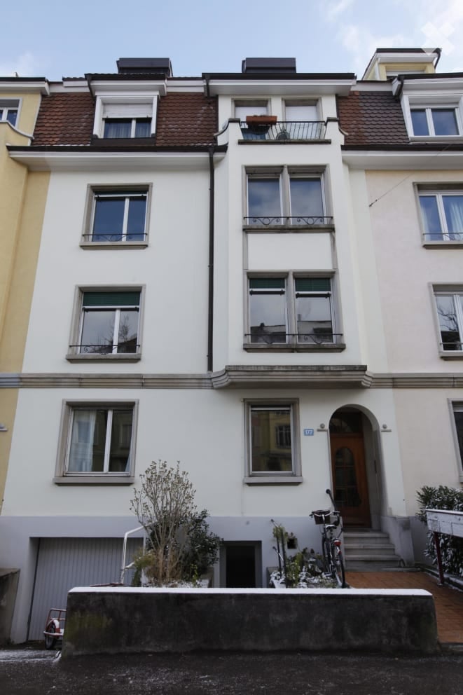 A multi-story residential building with white walls, multiple windows, and a brown tiled roof. Features include a garage entrance, small balcony, and a bicycle parked near the entrance.