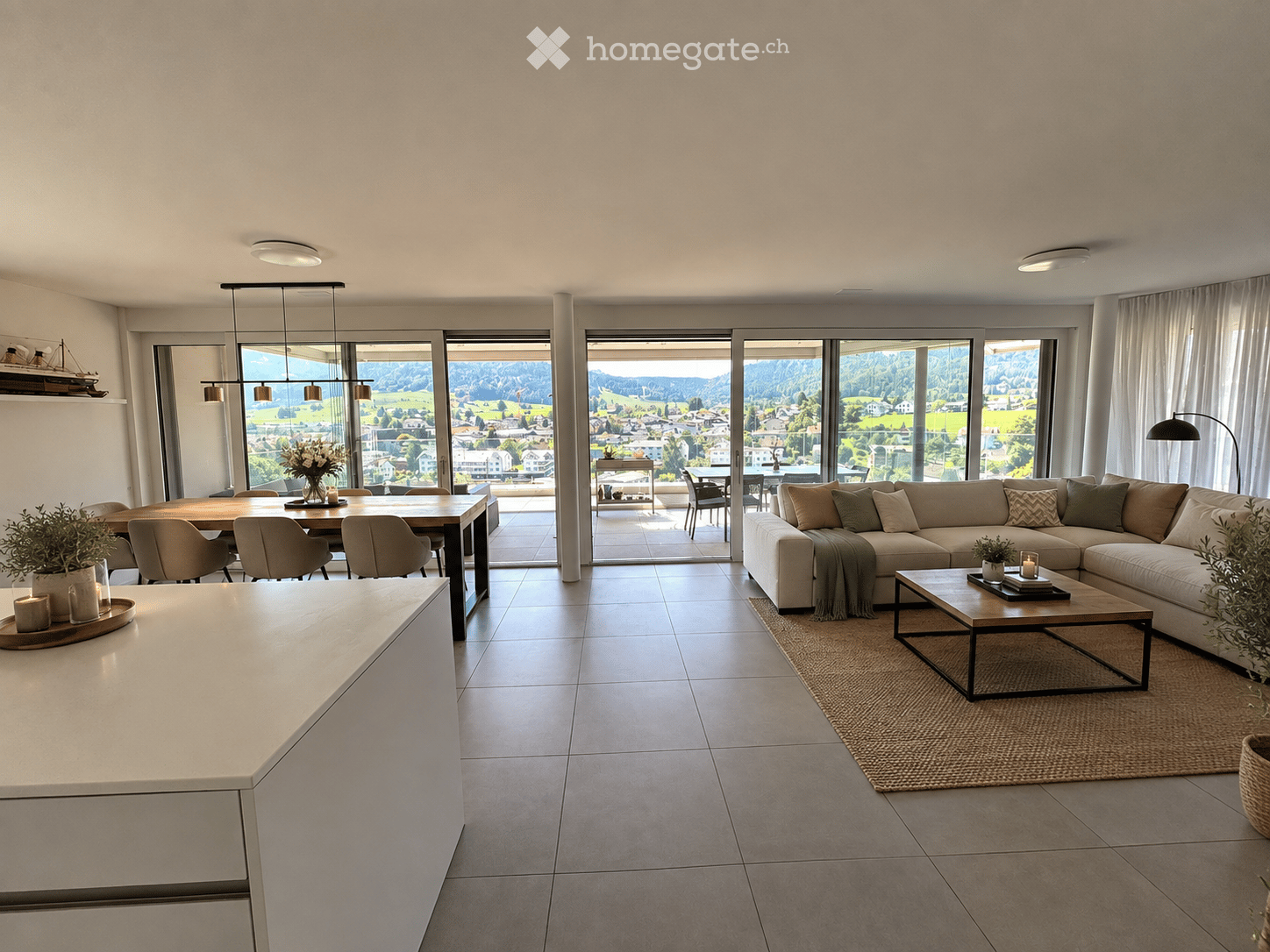 Modern open-plan living and dining area with large windows offering scenic views, featuring a sectional sofa, coffee table, dining table with chairs, and decorative elements like candles and plants.