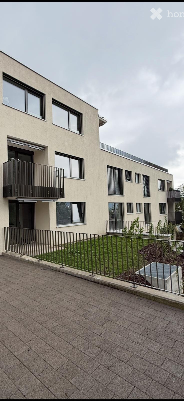 Modern multi-story building with large windows, balconies, and a paved ground-level area with a metal fence and small garden.