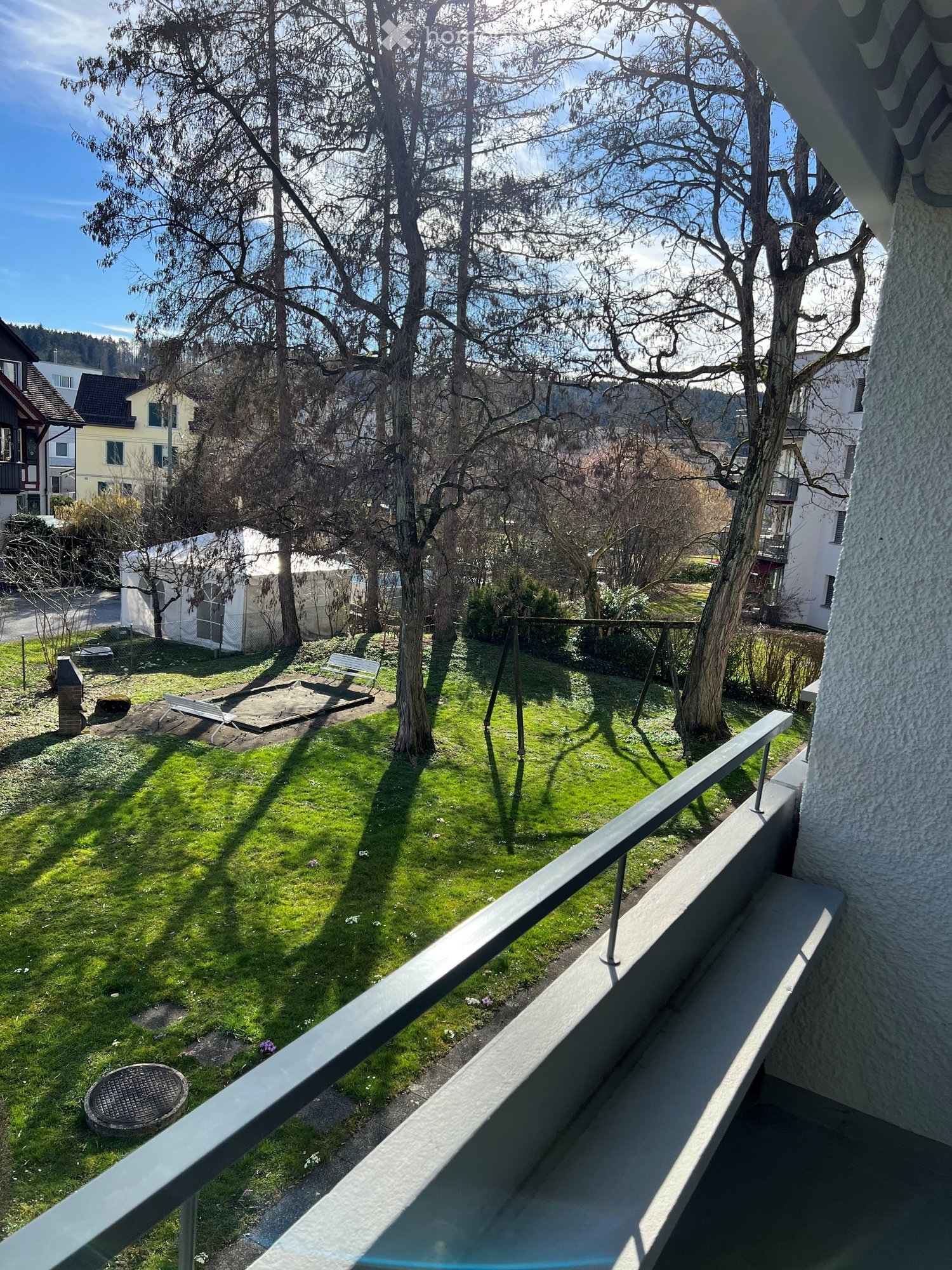 Balcony overlooking a grassy garden area with trees, a bench, and a small structure in the background.