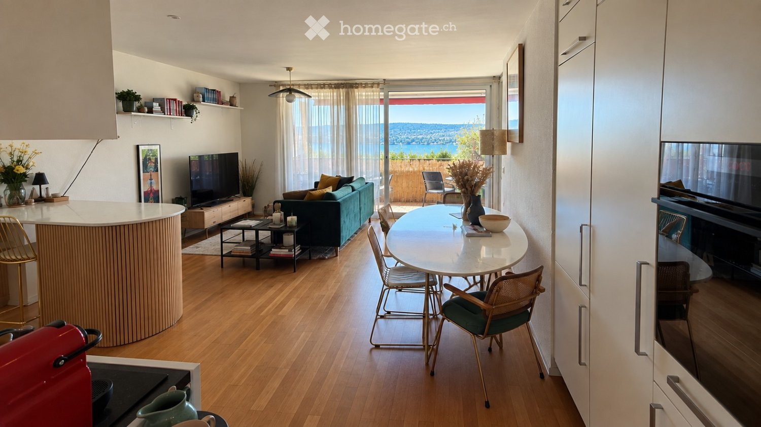 Modern open-plan living and dining area with wooden flooring, large windows offering a scenic view, a sofa, coffee table, dining table with chairs, and a kitchen island.