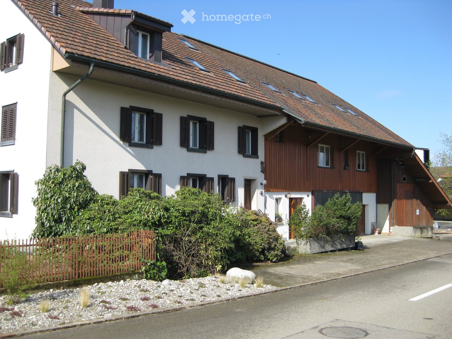 Two-story house with a red-tiled roof, white and brown exterior, multiple windows with shutters, and a small garden area with bushes and a fence.