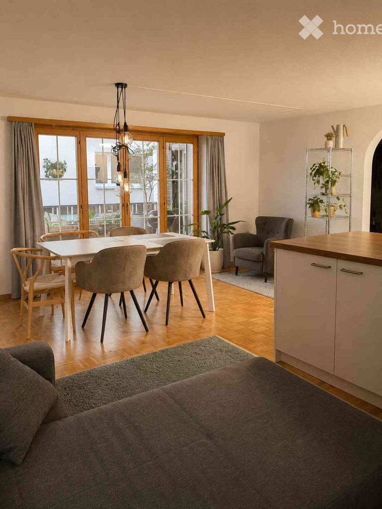 Modern open-plan living and dining area with a grey sofa, dining table with chairs, kitchen cabinetry, potted plants, and a shelf with decorative items.