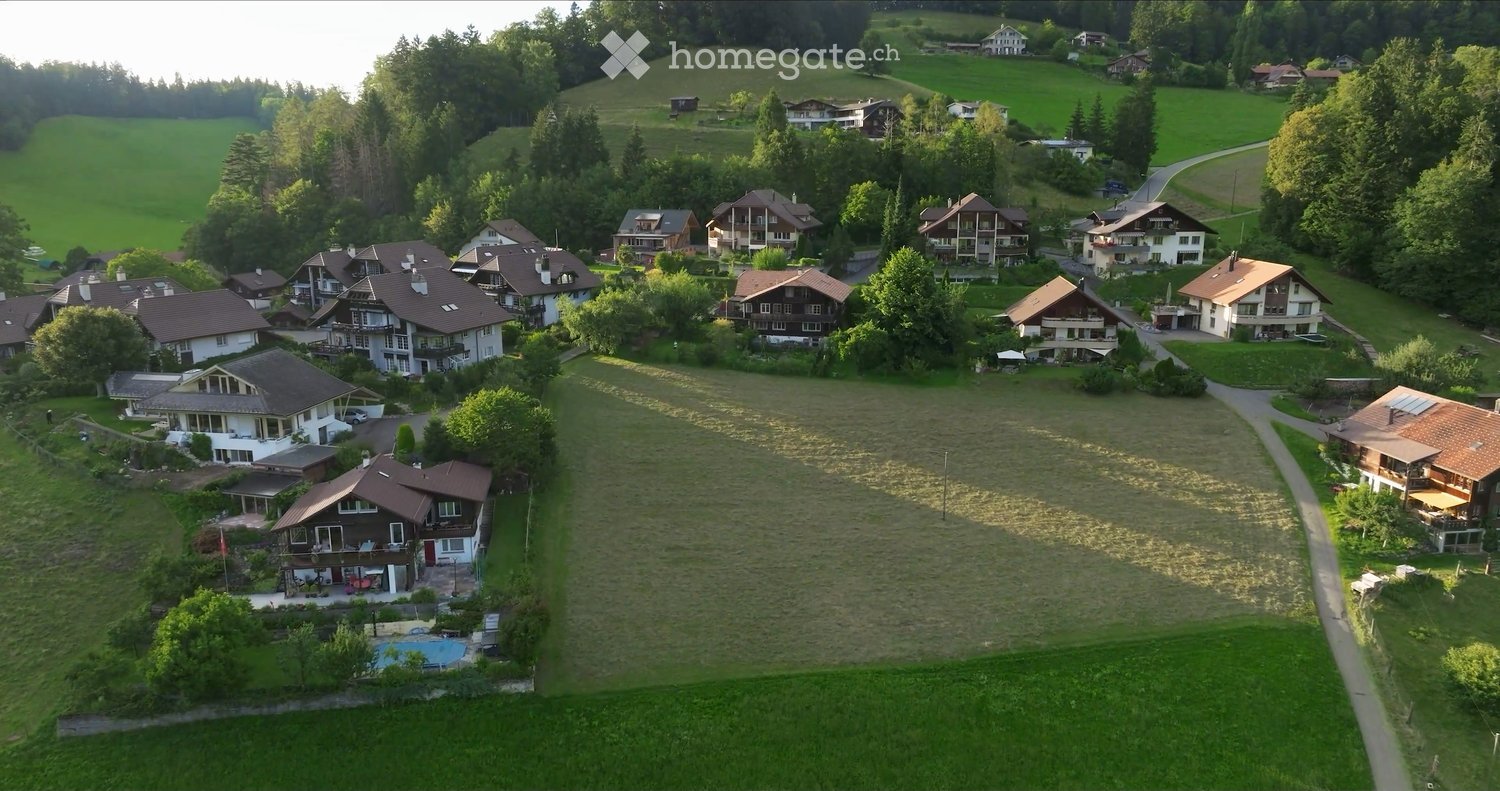 Aerial view of a residential area with multiple houses, surrounded by green fields and trees, with a swimming pool visible in the foreground.