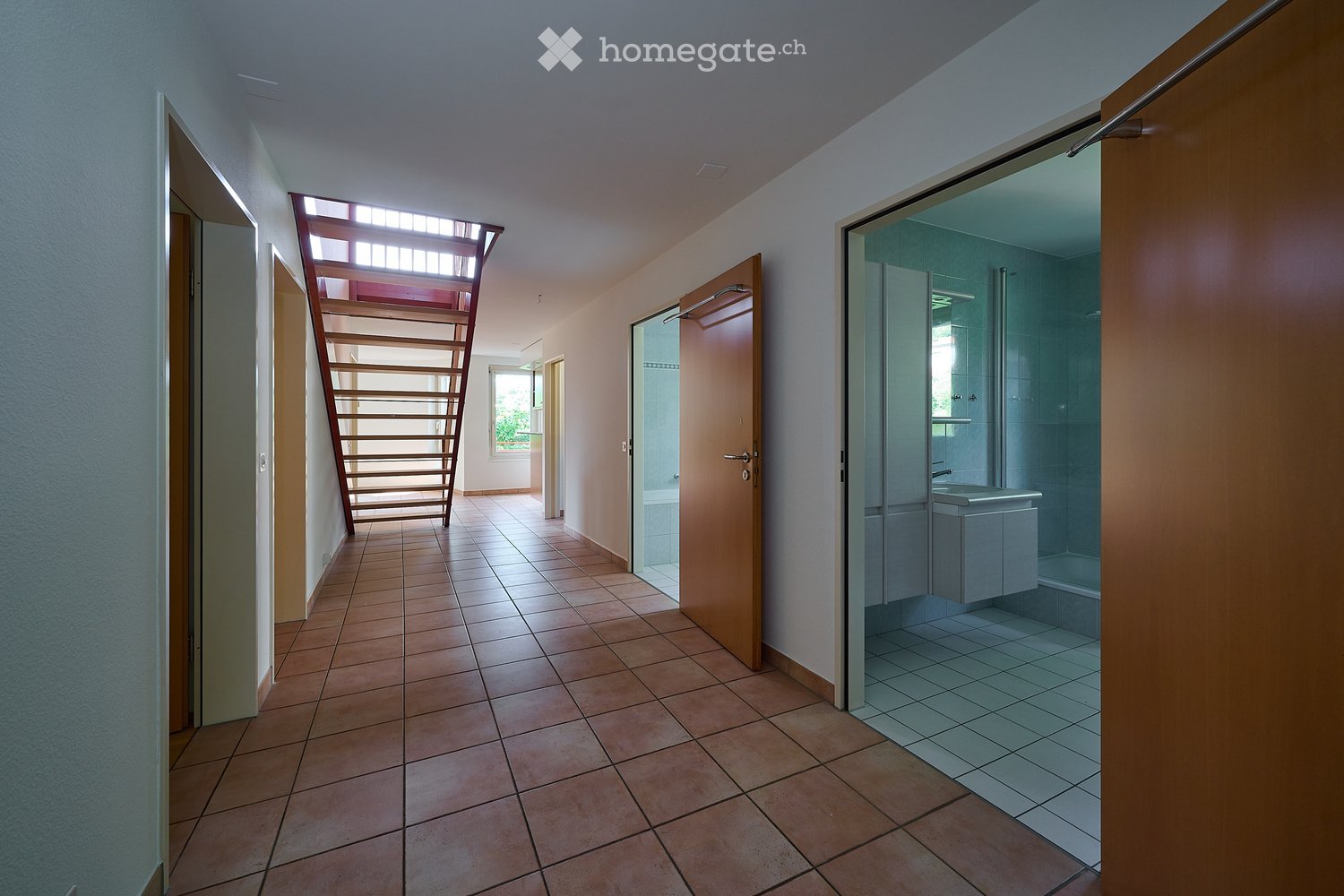 Empty hallway with tiled floor, wooden doors, and a staircase leading to an upper level. A bathroom is visible to the right with a sink and shower area.