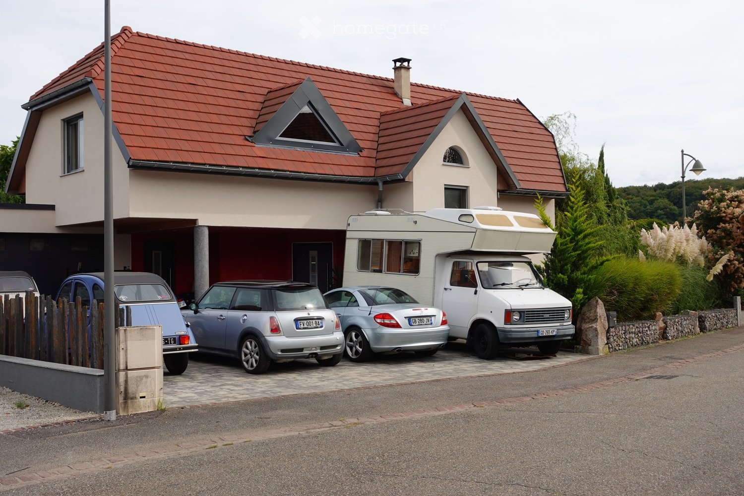 Two-story house with red-tiled roof, beige walls, and multiple windows. A camper van and several cars are parked in the driveway.