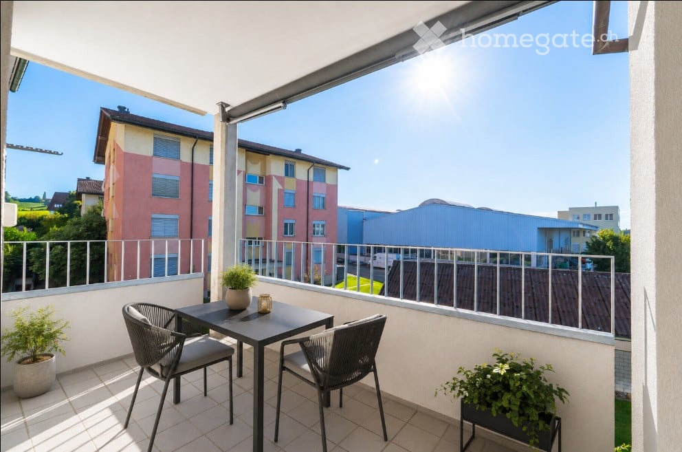Balcony with a small table and two chairs, potted plants, and a view of surrounding buildings and greenery.