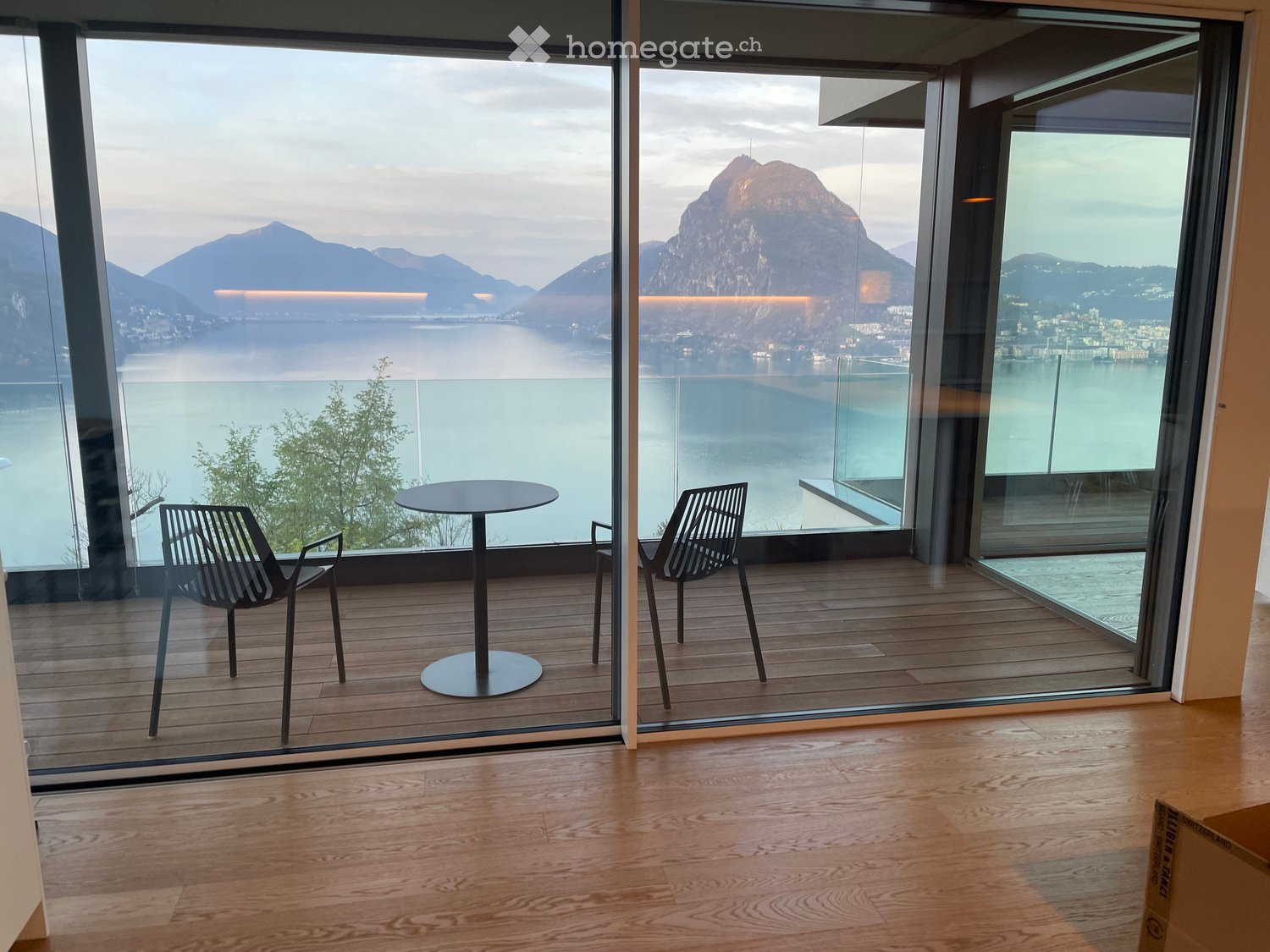 Modern balcony with wooden flooring, large glass walls offering a scenic view of a lake and mountains, equipped with a round table and two contemporary chairs.