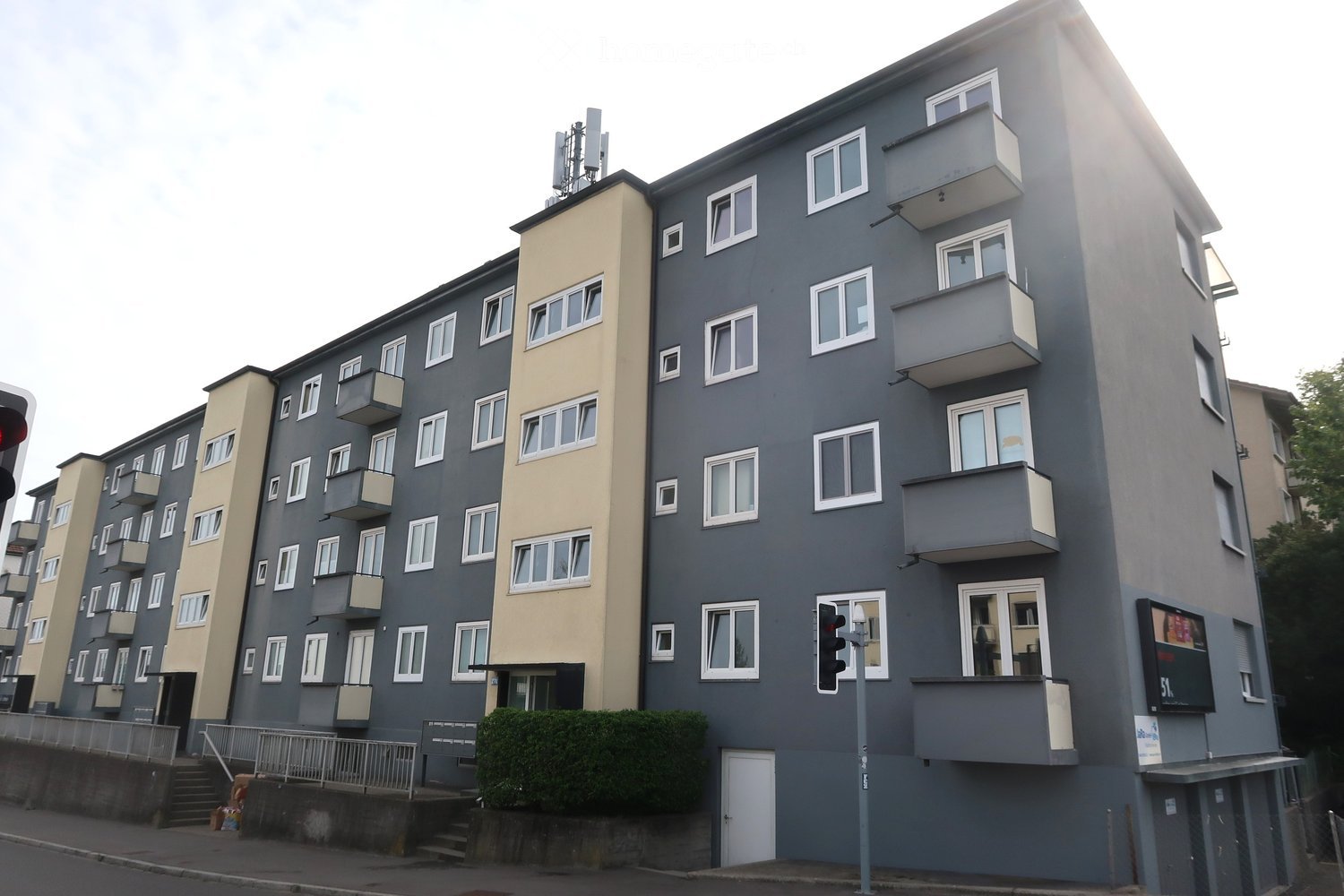 Multi-story apartment building with grey and beige facades, multiple windows, balconies, and a raised ground floor entrance.
