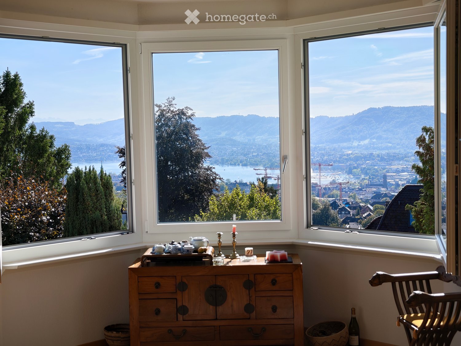 Corner room with large windows offering scenic views of mountains and water, furnished with a wooden dresser, teapot, candles, a wooden chair, and a bottle on the floor.