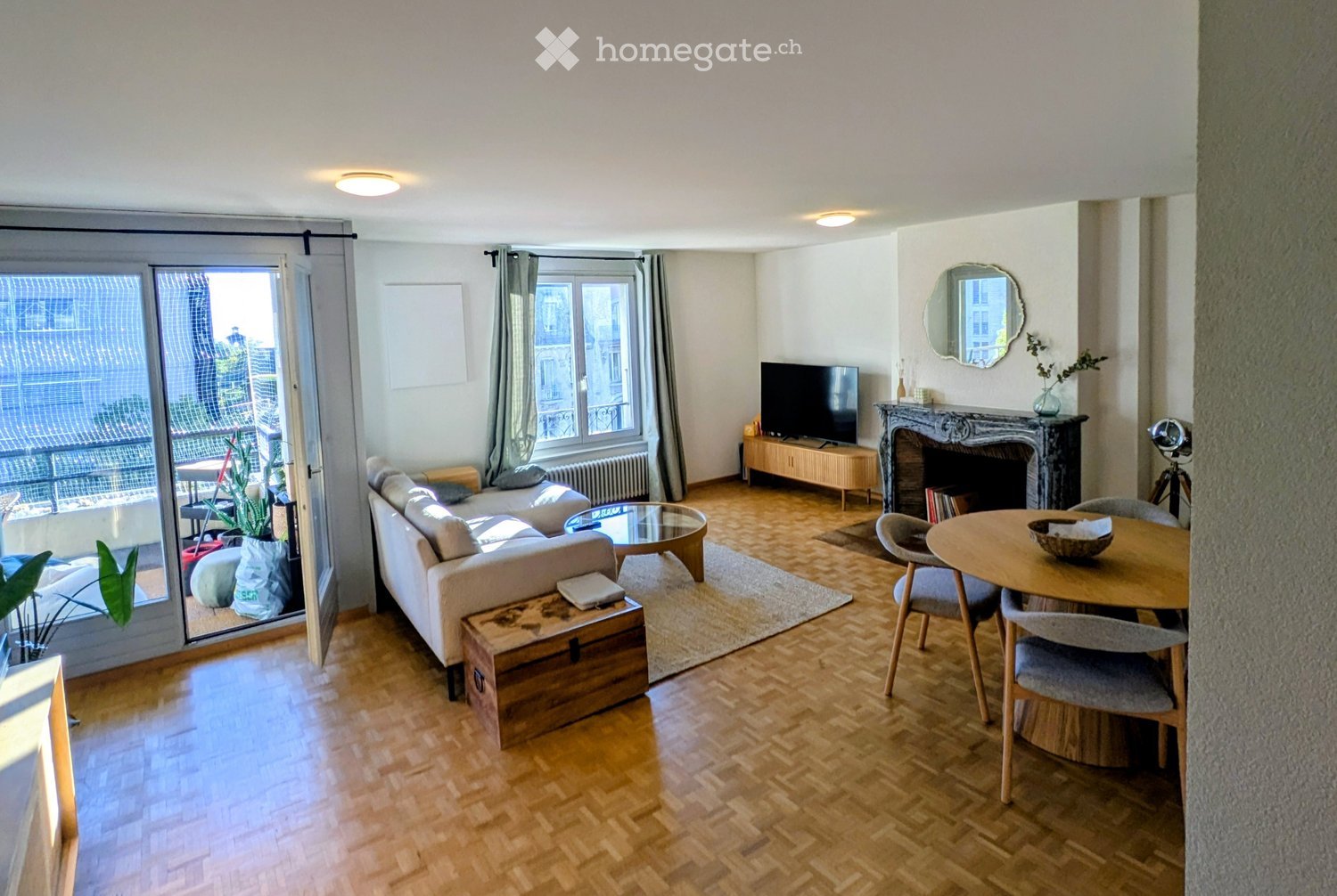 Bright living room with wooden flooring, large windows, balcony access, sofa, coffee table, dining table, fireplace, TV, and decorative items.