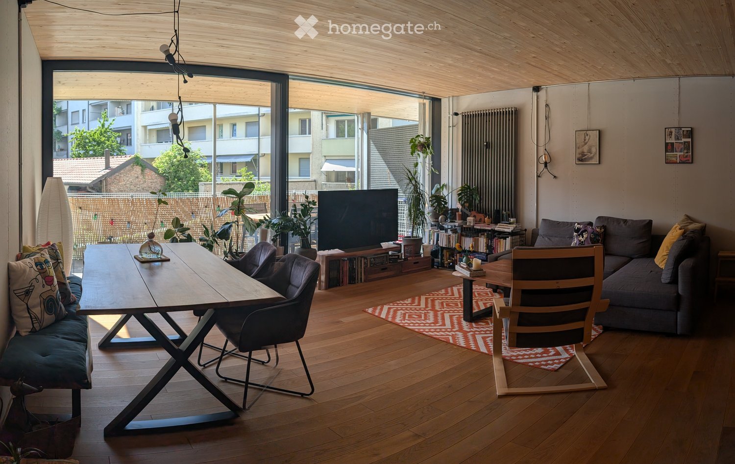 Modern living room with wooden flooring, large windows, a grey sofa, a wooden dining table, chairs, a television, and decorative plants.