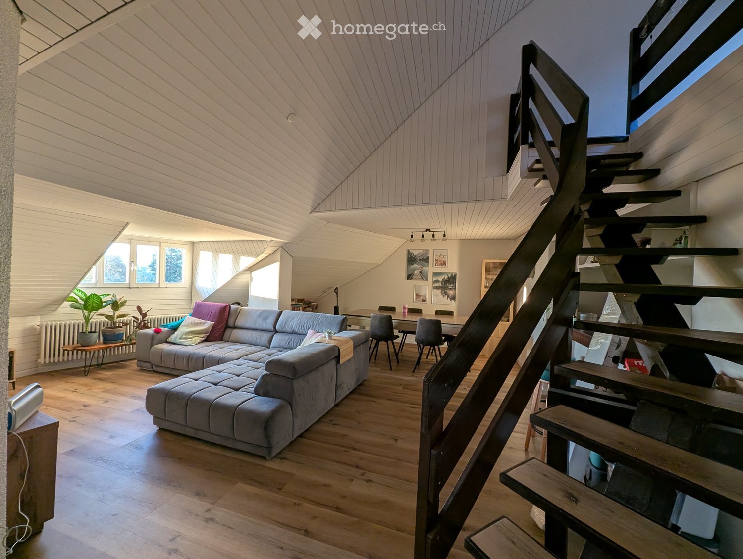 Open-plan living and dining area with wooden flooring, grey sectional sofa, coffee table, dining table with chairs, and a staircase leading to an upper level.