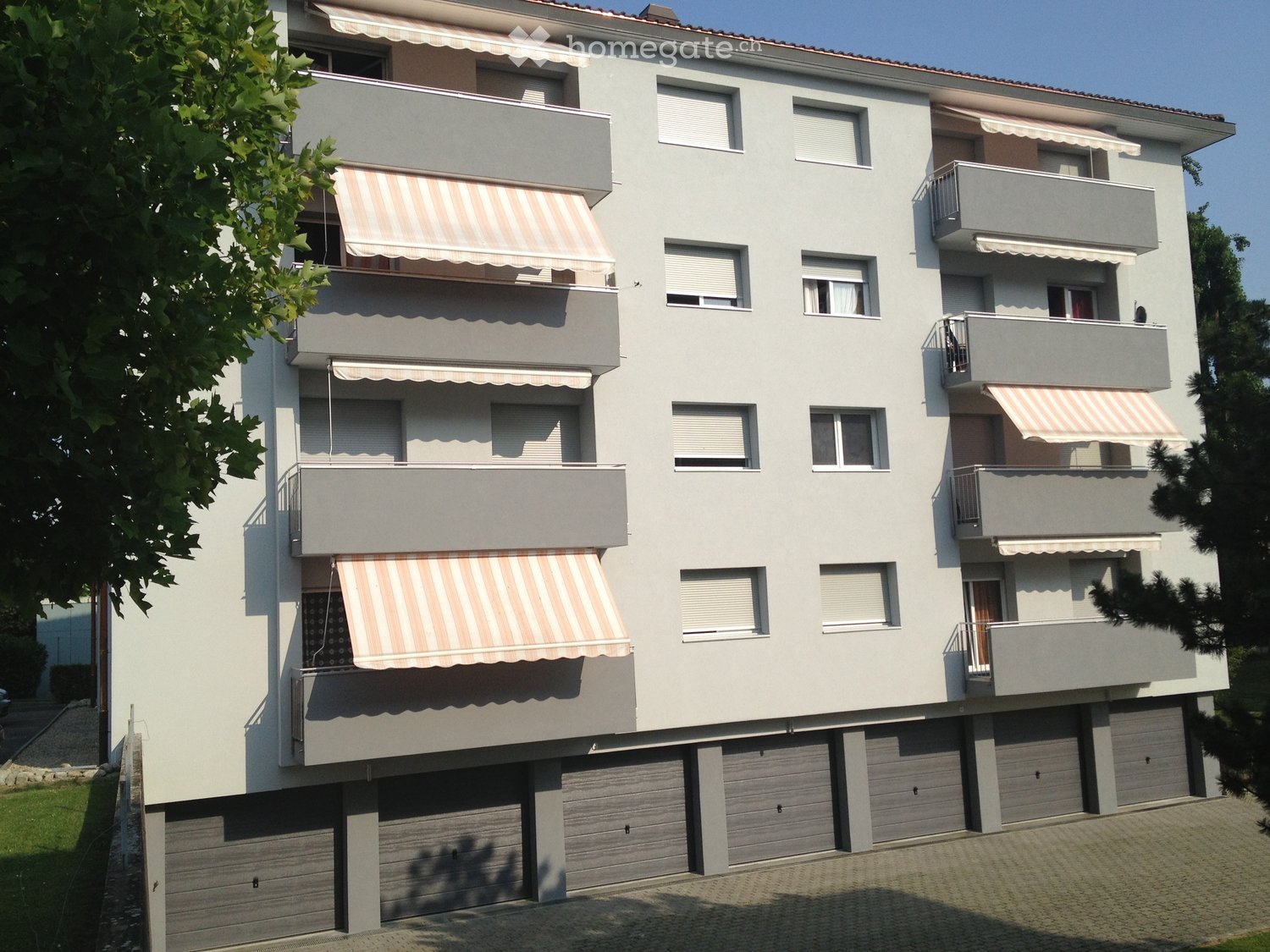 Four-story residential building with multiple balconies, striped awnings, and a row of garages on the ground floor.