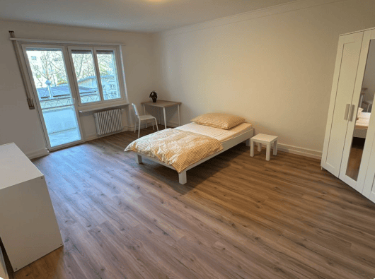 Bright bedroom with wooden flooring, large window leading to a balcony, bed with pillows and blanket, nightstand, chair, small table, and wardrobe.