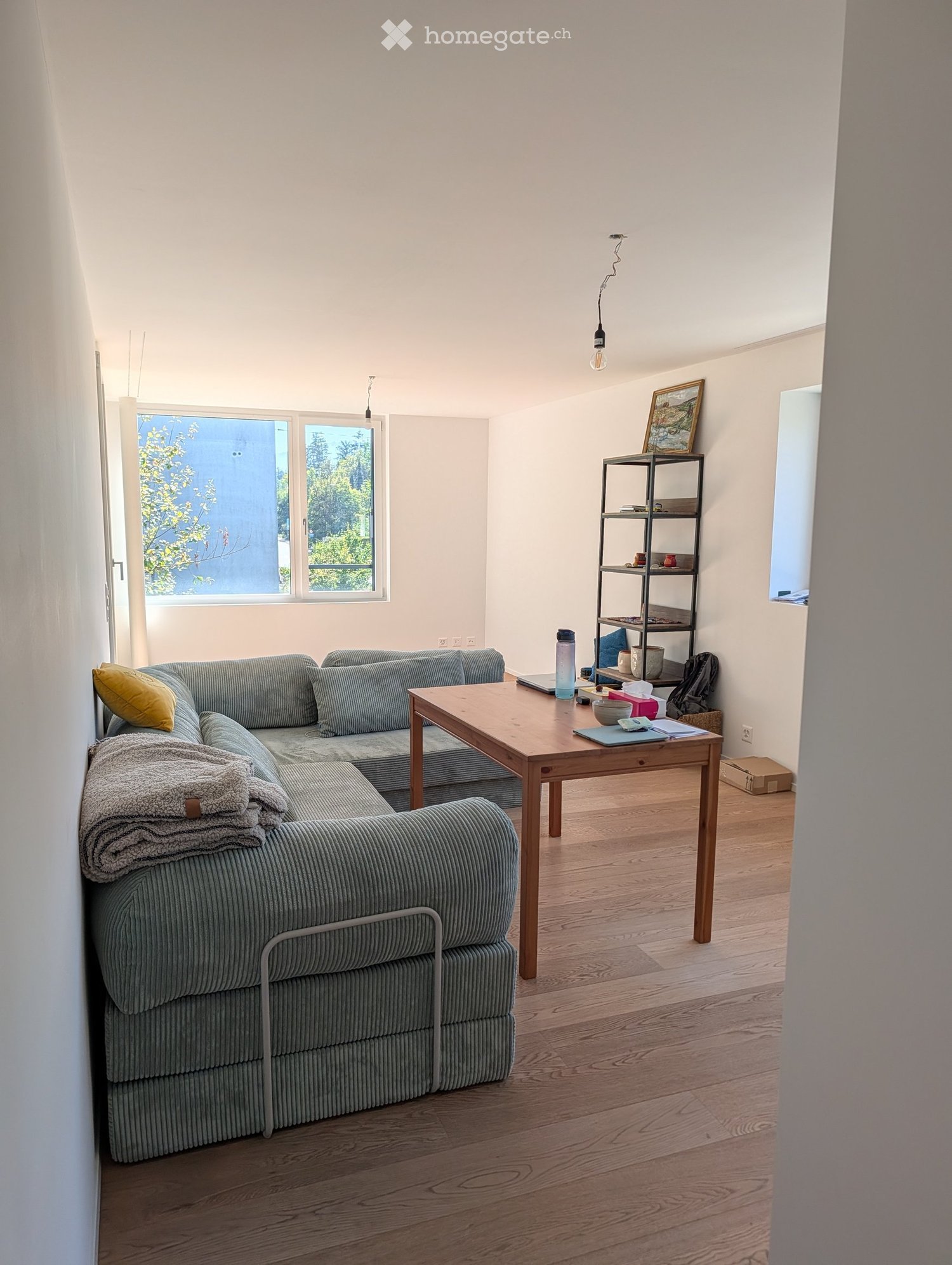 Living room with a grey sectional sofa, wooden coffee table, shelf with books and decorative items, and large windows with a view of greenery.