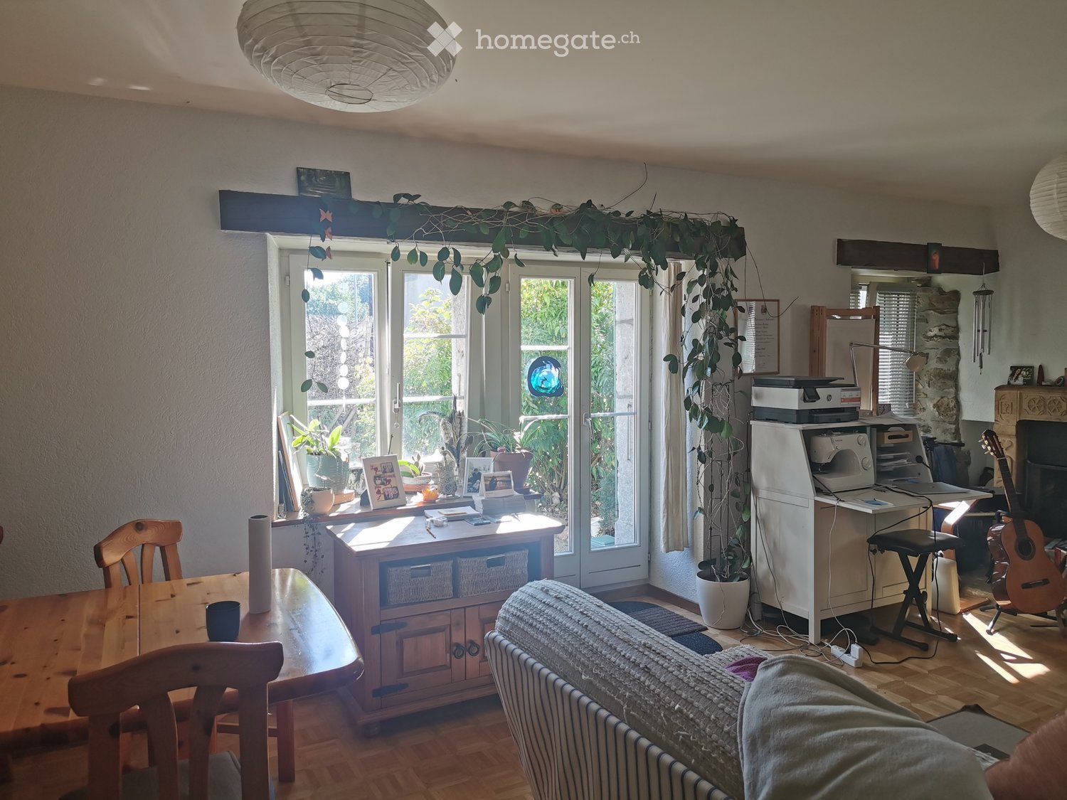 Bright living space with a sofa, dining table, chairs, guitar, printer, and decorative plants. Large windows and a balcony door provide natural light.