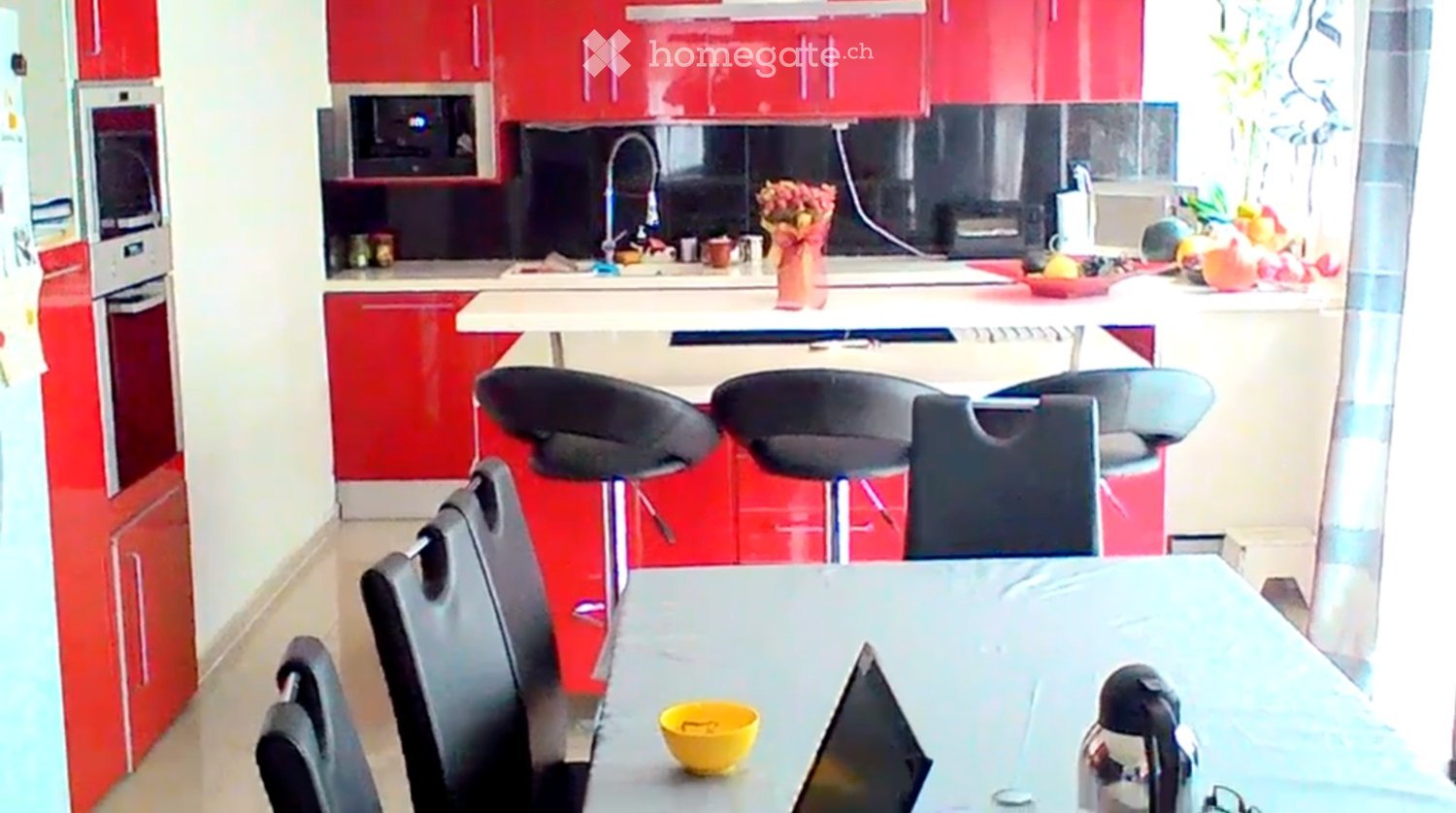 Modern kitchen with red cabinets, white countertops, and stainless steel appliances. Includes a dining area with a table, chairs, and stools.