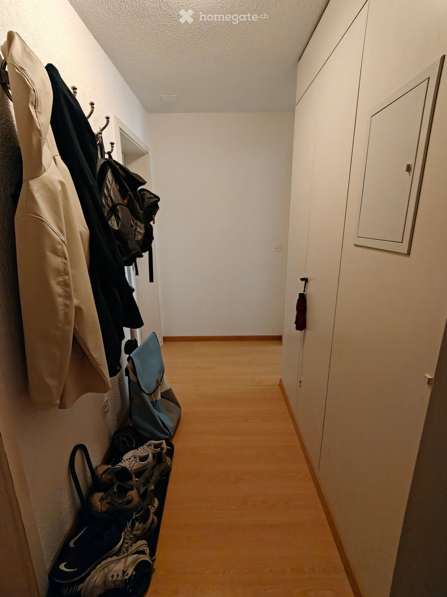 A narrow hallway with wooden flooring, white walls, and a coat rack holding jackets and bags. A closet door is visible on the right side.