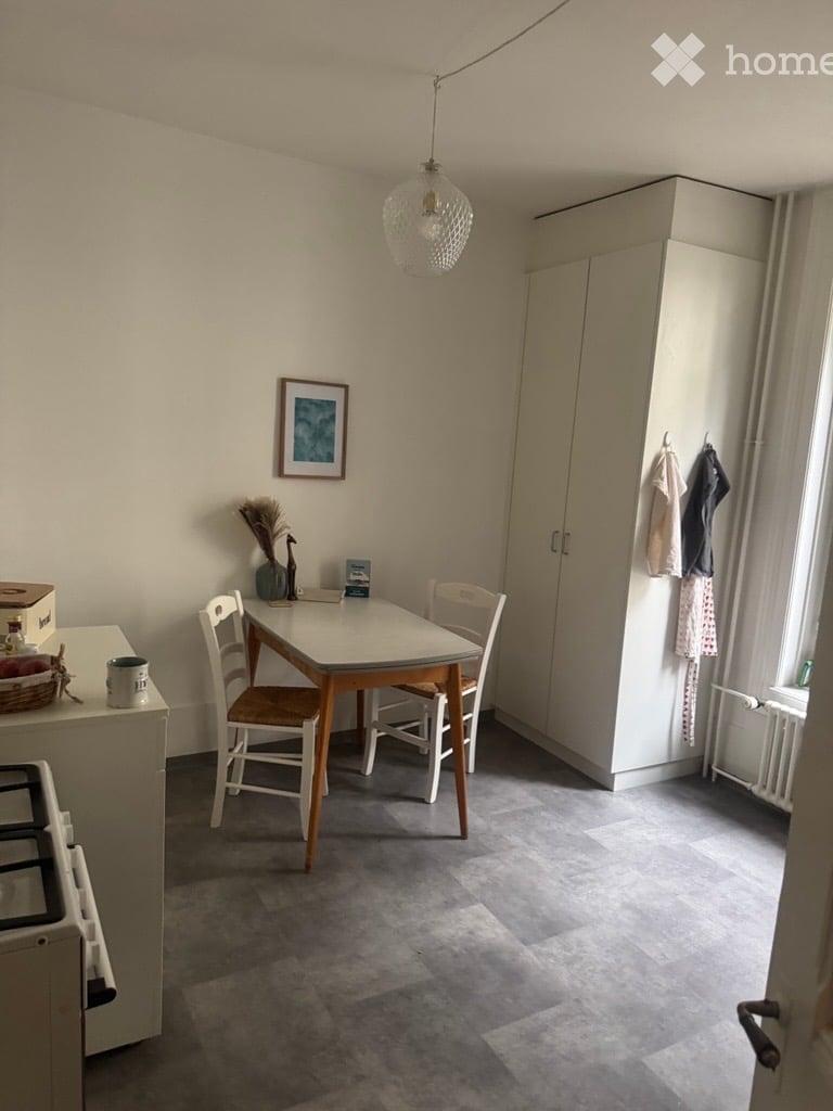A small, furnished room with a dining table, chairs, a white cupboard, a picture frame on the wall, and a hanging light fixture.