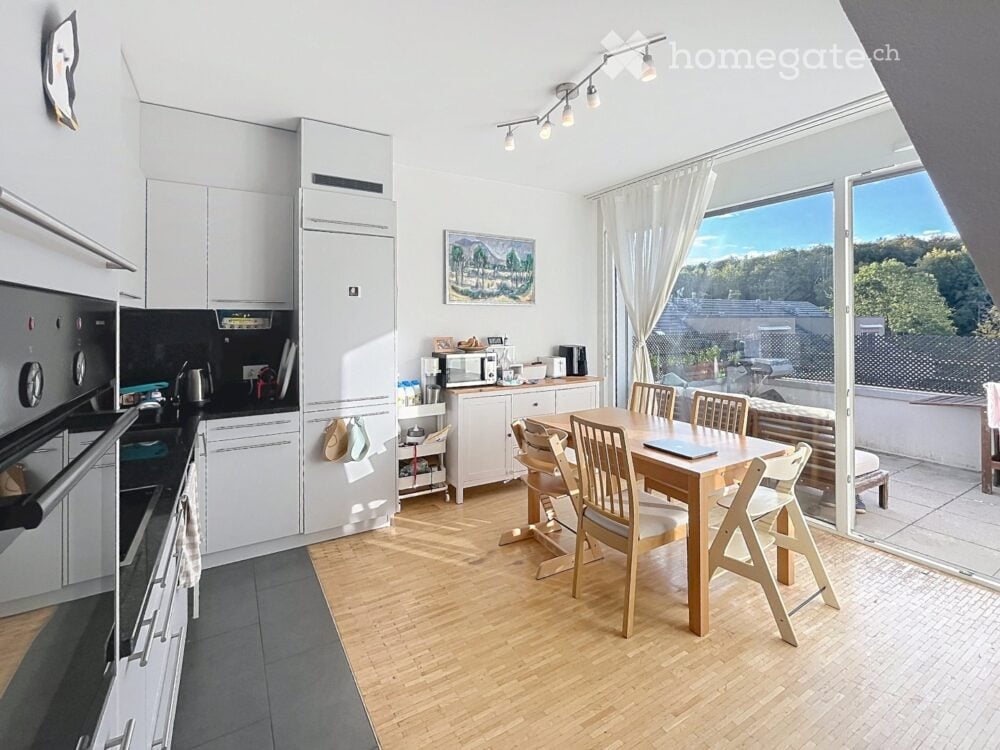 Open-plan kitchen and dining area with white cabinets, wooden flooring, and large windows offering a scenic view. Includes a dining table with chairs, a laptop, and various kitchen appliances.