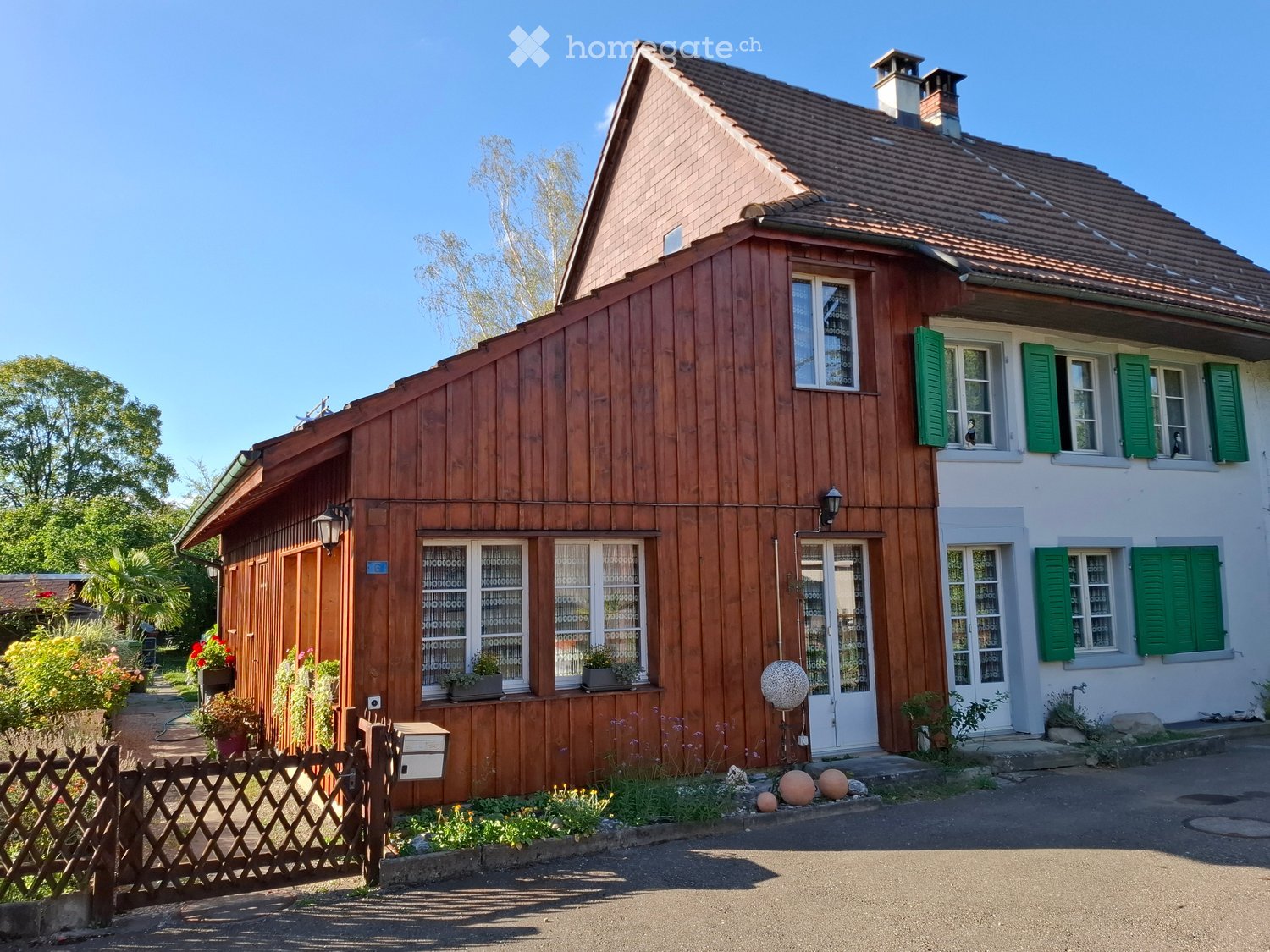 Two-story house with wooden siding, green shutters, and a tiled roof. Features a small garden with plants and a wooden fence.