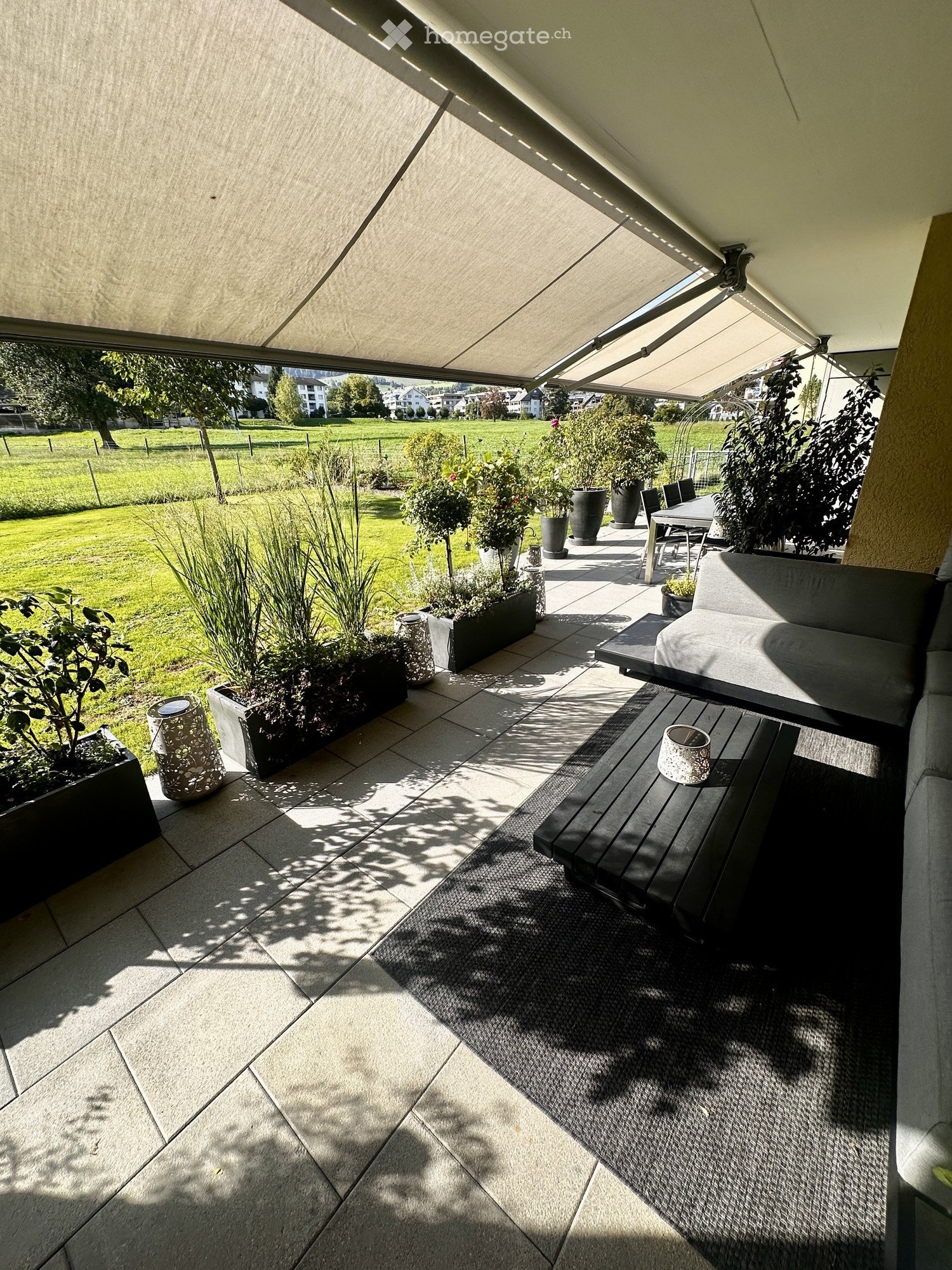 A modern balcony with a retractable awning, tiled floor, and outdoor furniture including a sofa, table, chairs, and a bench. Potted plants and a view of a green lawn are visible.