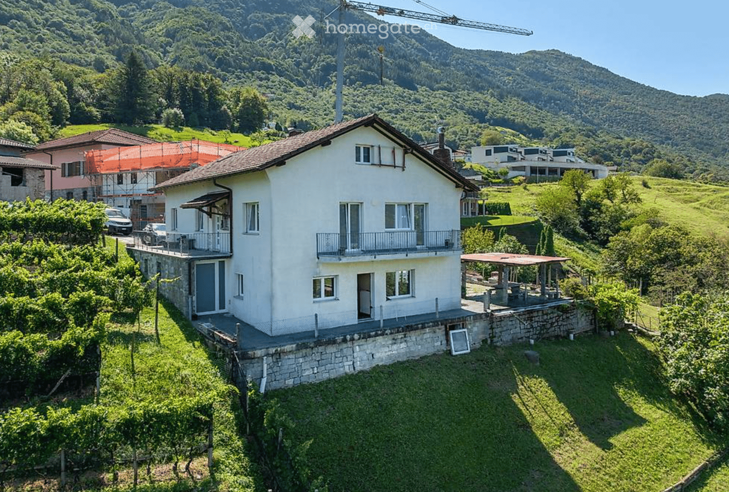 Two-story white house with balconies, surrounded by greenery and vineyards, with a scenic mountain backdrop.