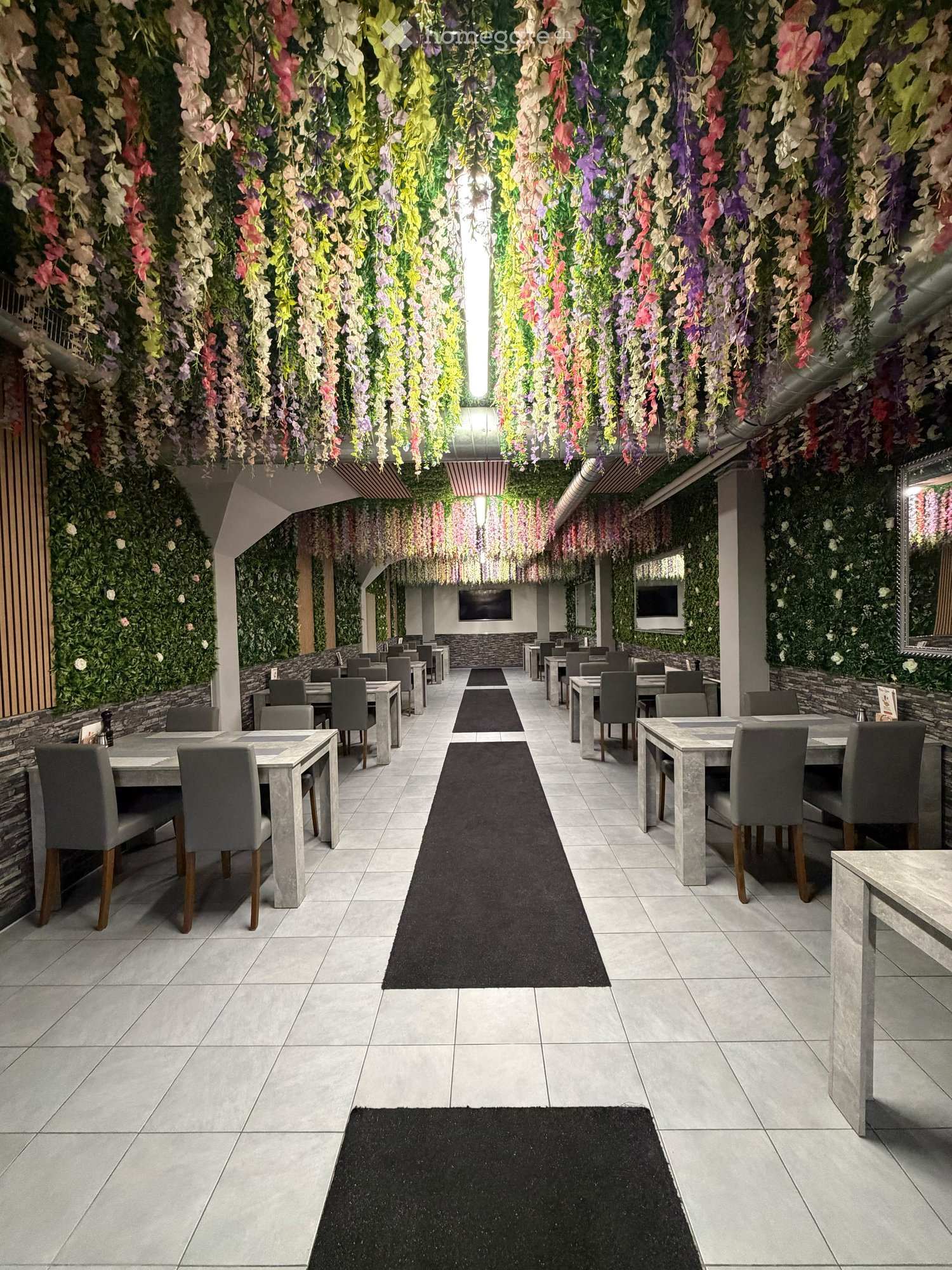 A modern restaurant with a long central aisle, multiple tables and chairs, and a floral vertical garden on the walls.