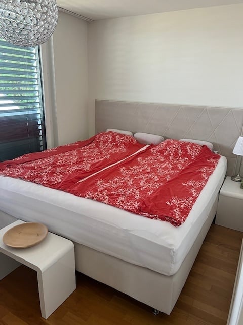 Modern bedroom with a large double bed, red patterned duvet, white nightstand with a lamp, and a small wooden stool with a decorative plate on it.