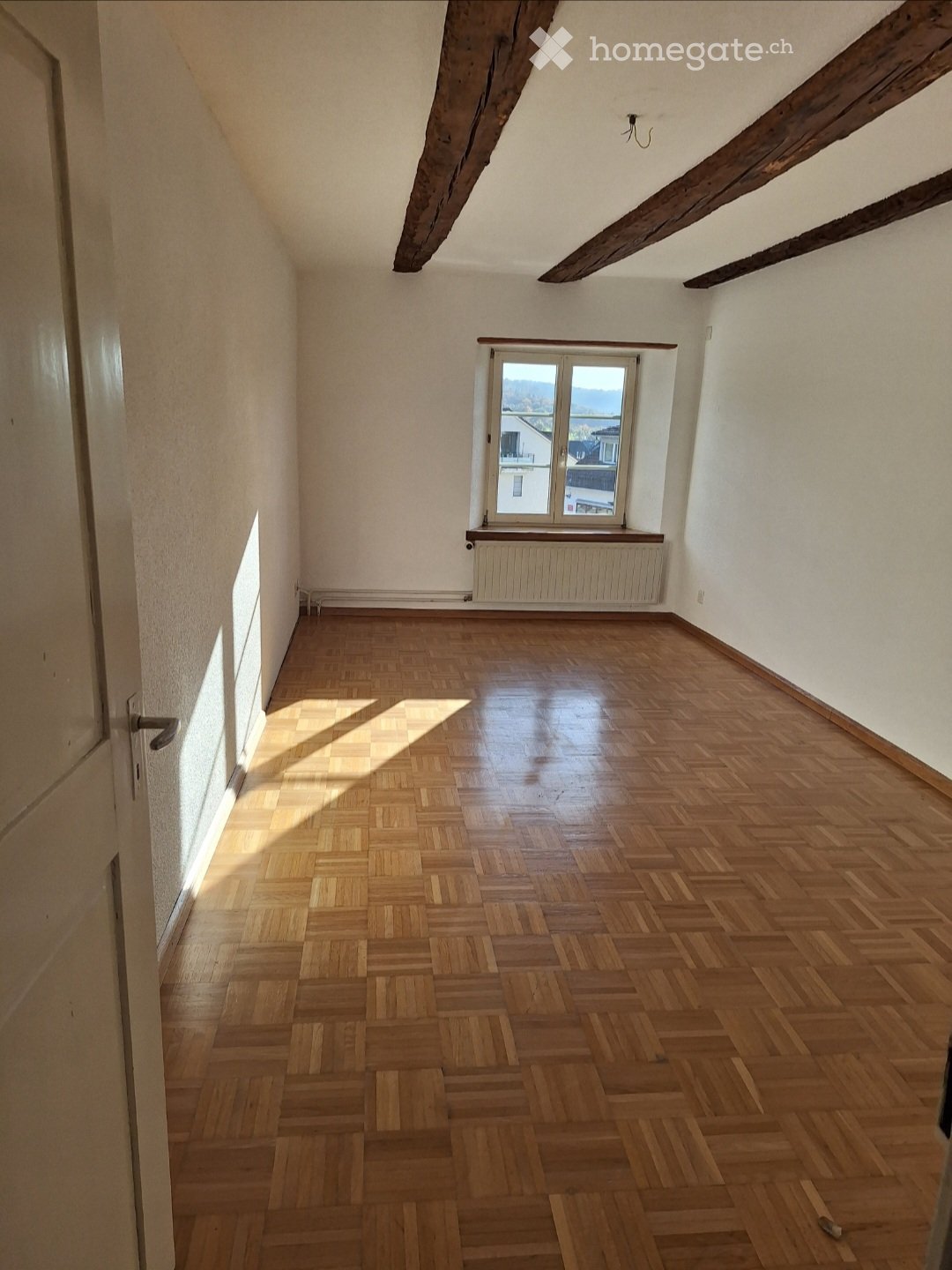 Empty room with wooden parquet flooring, white walls, exposed wooden beams on the ceiling, and a single window with a radiator below it.