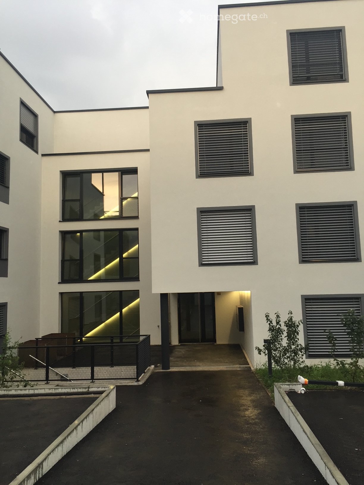 Modern two-story building with white walls, large windows, and a raised ground floor entrance. The building features a sleek design with black-framed windows and a small garden area in front.