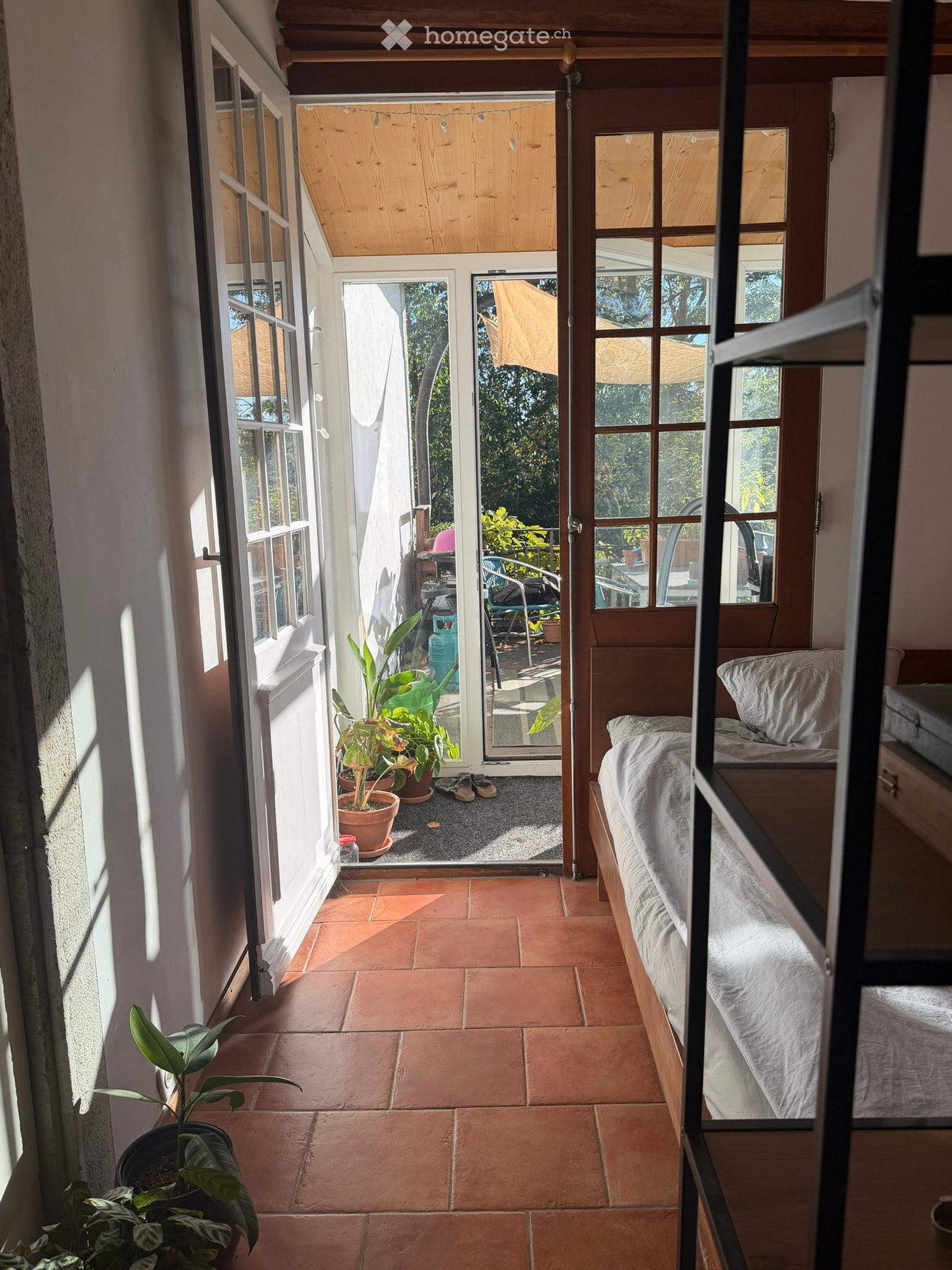 Cozy bedroom with terracotta tile floor, bed with white bedding, nightstand, and potted plants near the window and door leading to outdoor area.