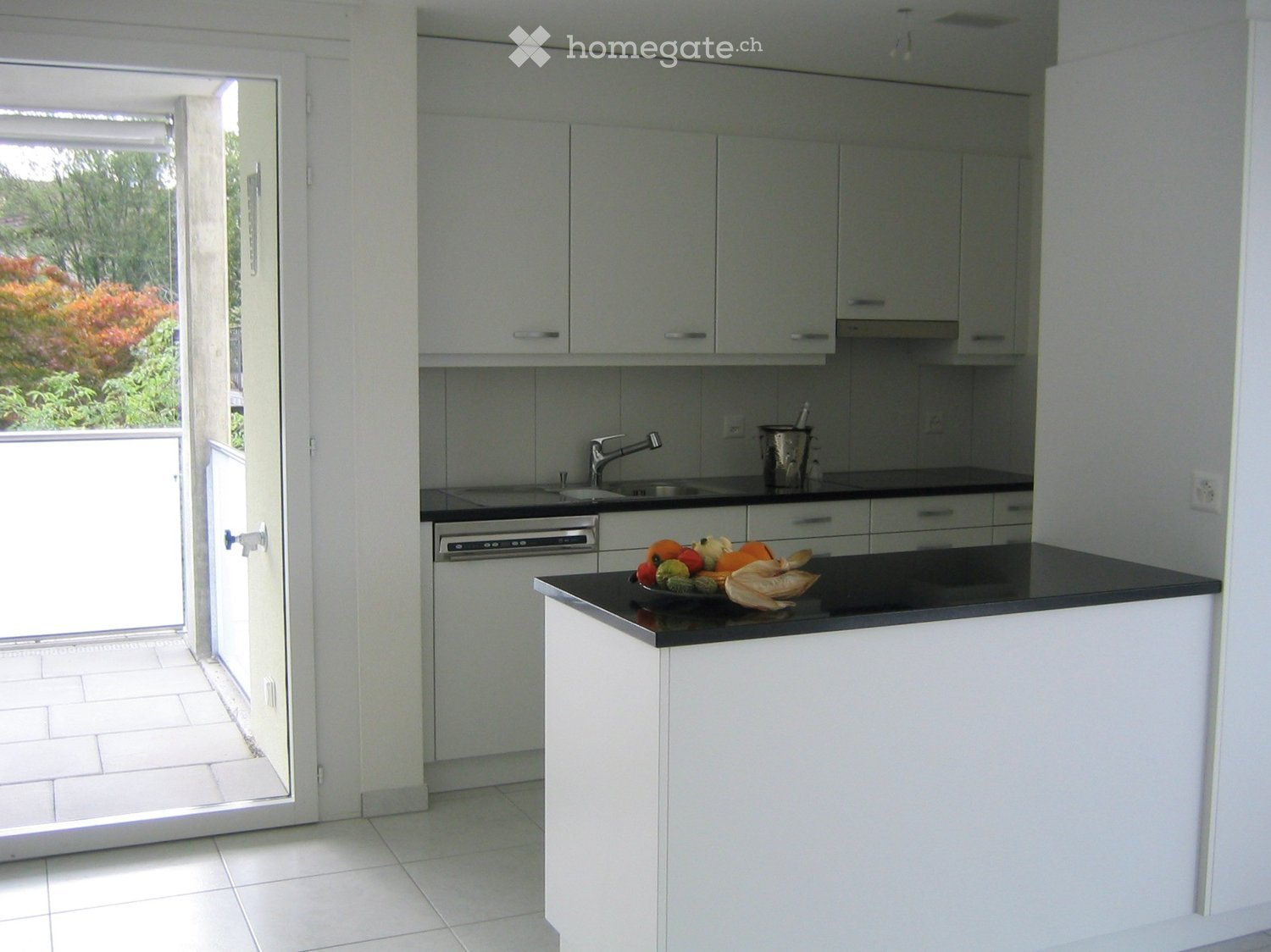 Modern kitchen with white cabinets, black countertops, a sink, a dishwasher, and a bowl of fruit on the counter. The kitchen has a sliding glass door leading to an outdoor area.