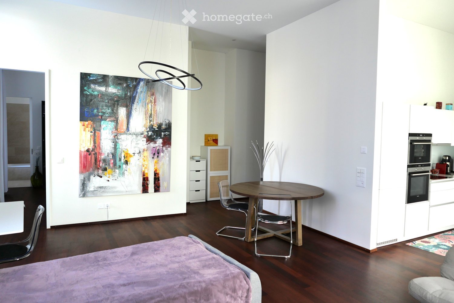 A modern, open-concept living space with a bed, dining table, chairs, a cabinet, a painting, and a small kitchen area with an oven.